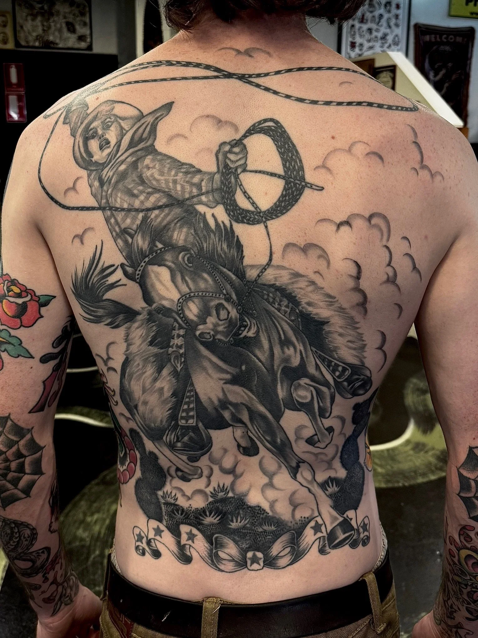 Black and gray tattoo on a man's back depicting a cowboy riding a bucking bull, with a lasso in hand, against a background of stylized clouds, made at Thanks Tattoo