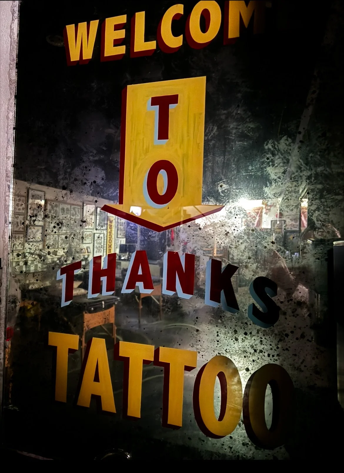 A glass door with a colorful sign that says 'Welcome To Thanks TATTOO' in bold red and yellow letters. The sign is displayed on the glass, which has some smudges and dirt.