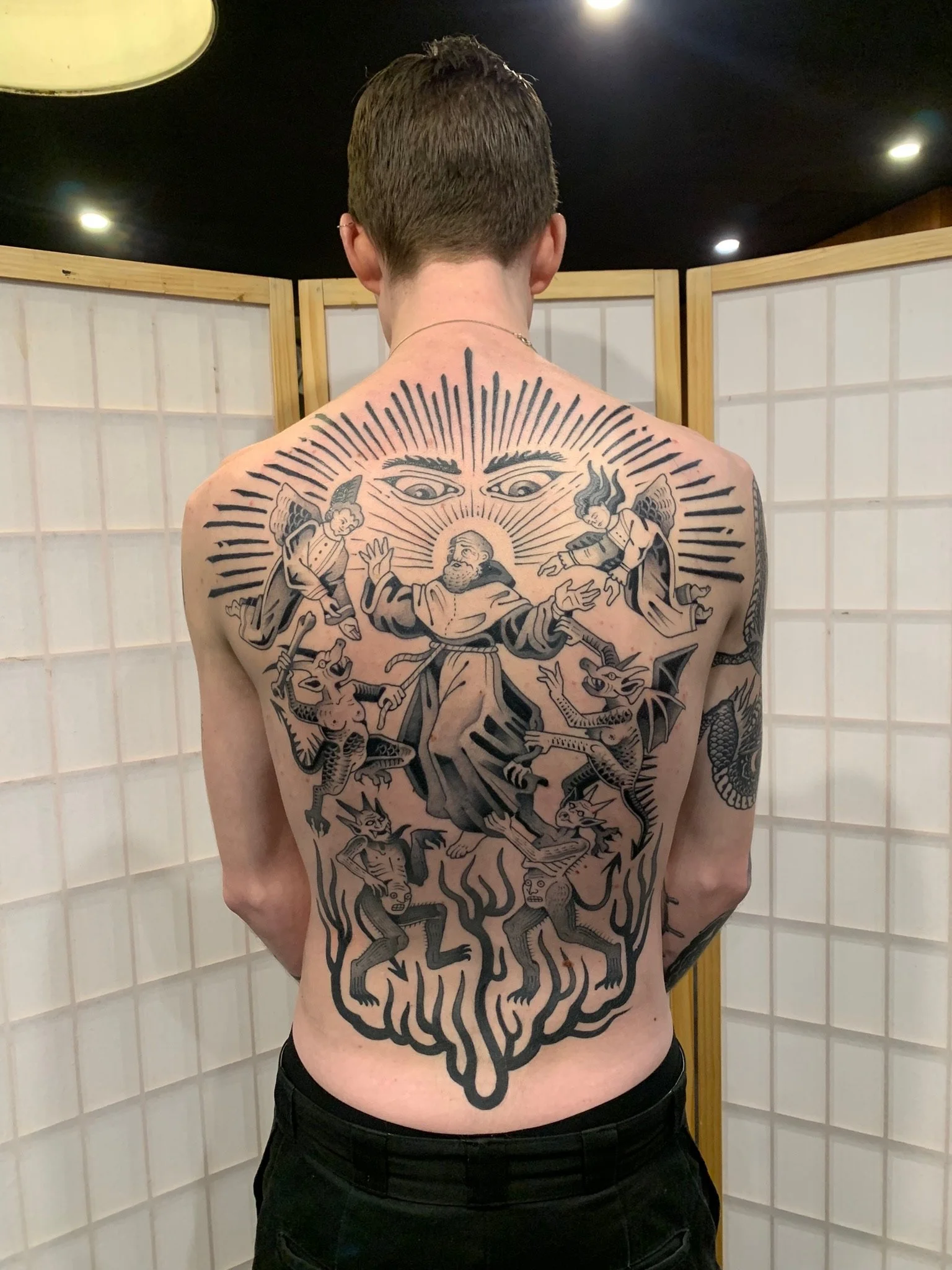 large, detailed black tattoo on their back depicting religious and mythical figures, including angels, demons, and a figure resembling a saint or biblical character, with upper eye-like symbols and radiant lines above, made at Thanks Tattoo