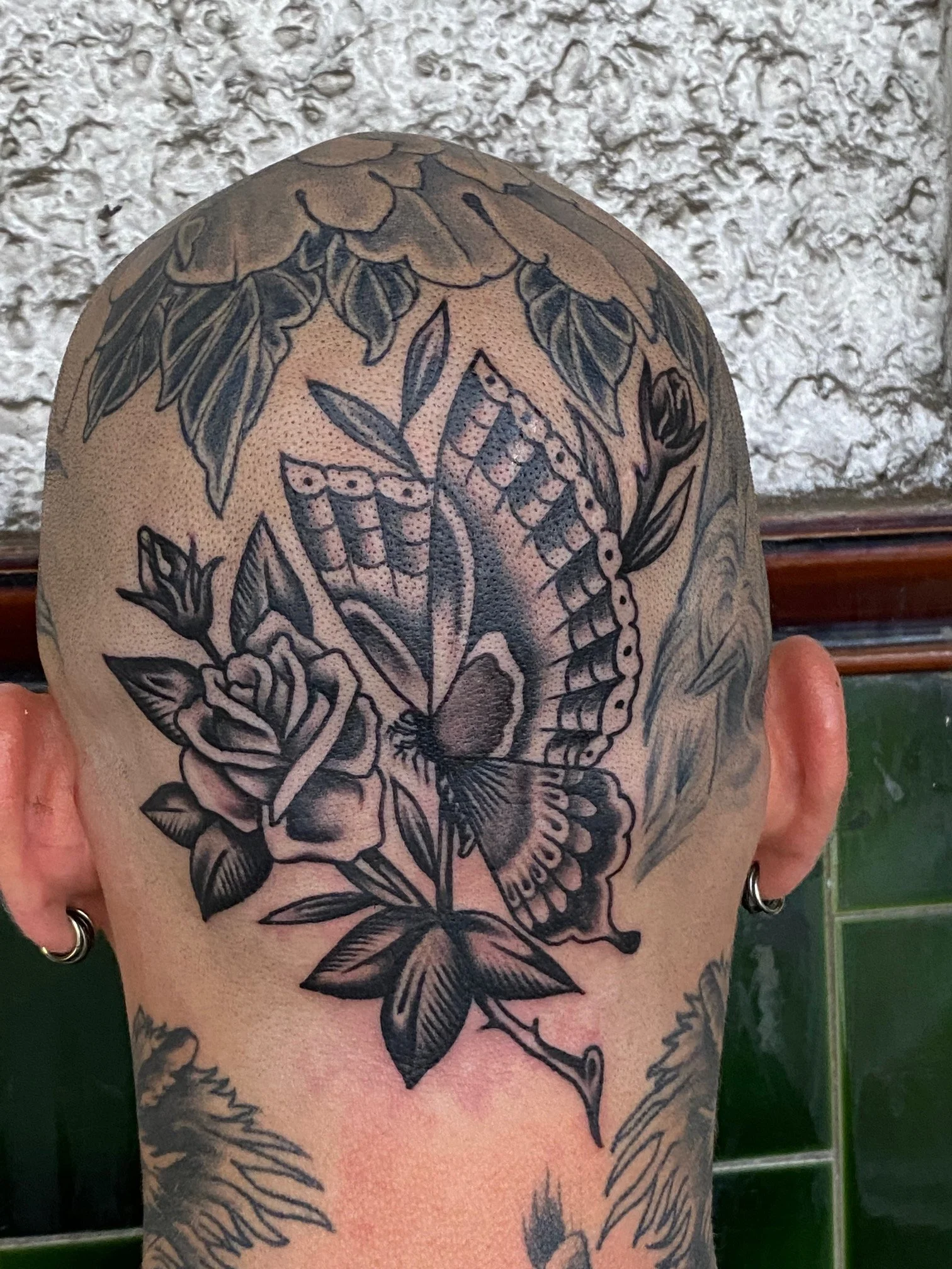 A black tattoo on the back of a person's head depicting a butterfly, flowers, and leaves against a textured background, made at Thanks Tattoo
