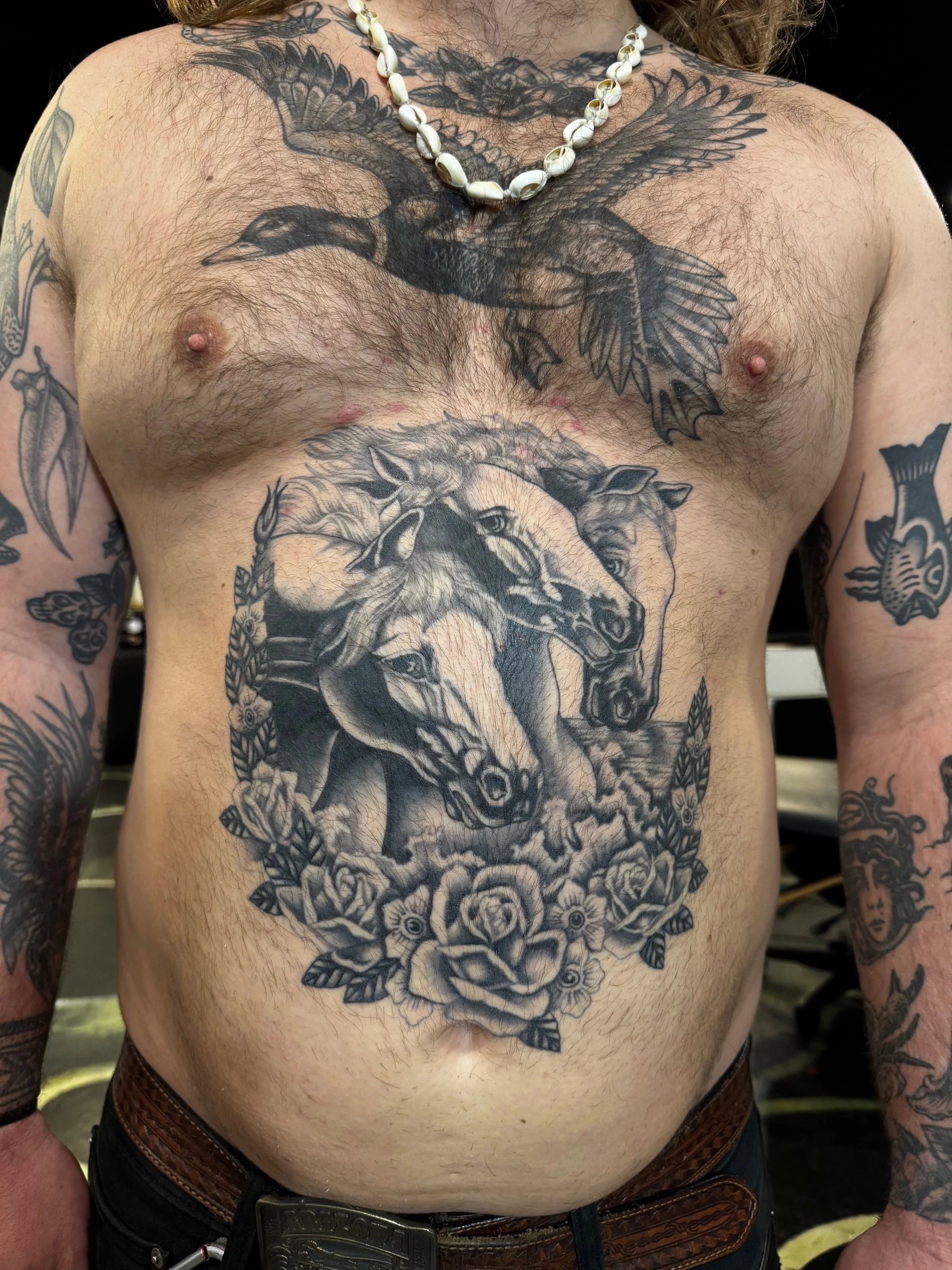 large tattoo of two horses' heads surrounded by flowers on their chest, a bird tattoo on their upper chest, and various tattoos on their arms. They are wearing a shell necklace and a belt with a large buckle, made at Thanks Tattoo
