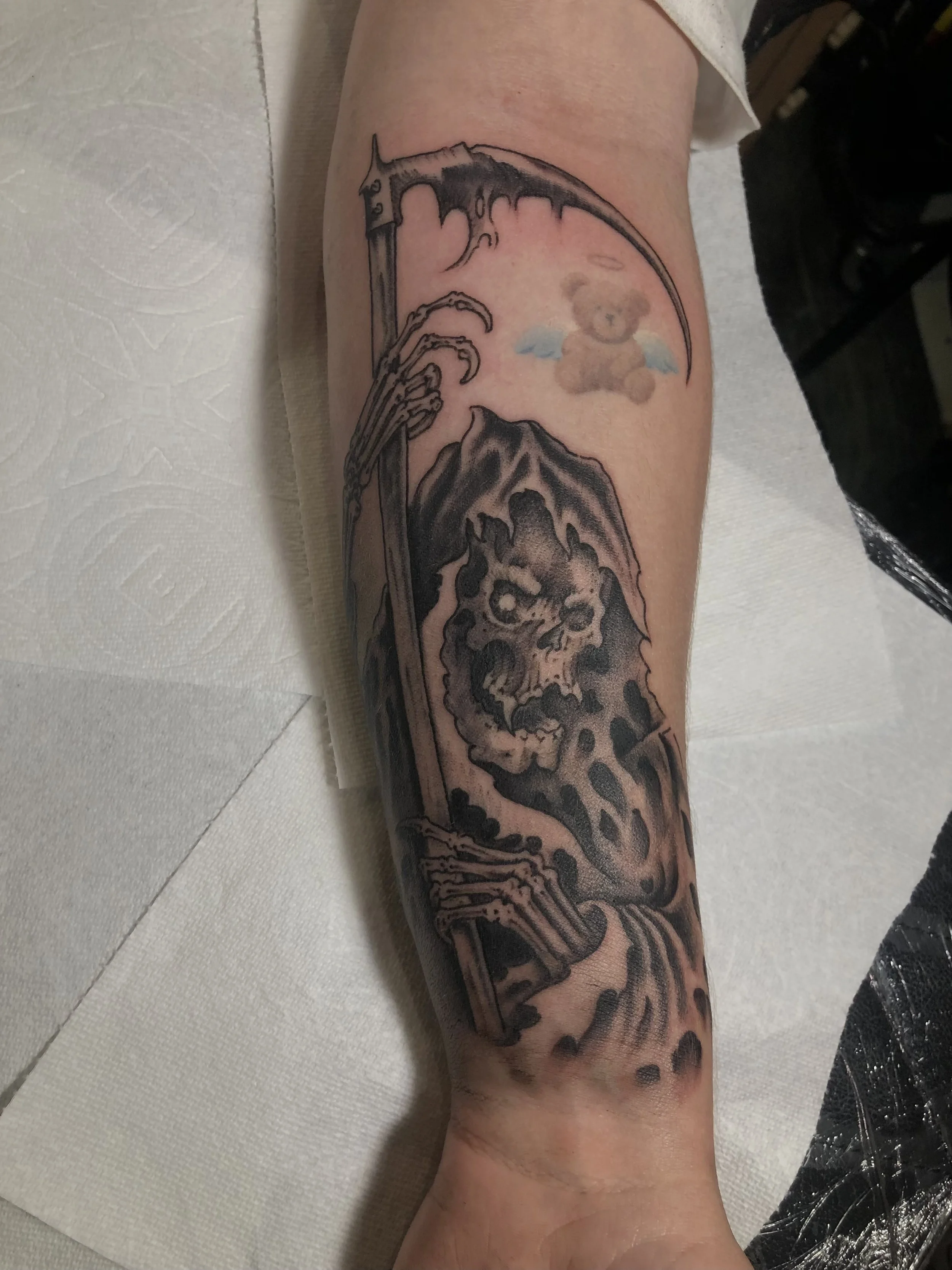 Black and gray tattoo of a Grim Reaper holding a scythe on a person's forearm, with a small cartoon teddy bear with blue wings and a halo above the Grim Reaper, made at Thanks Tattoo