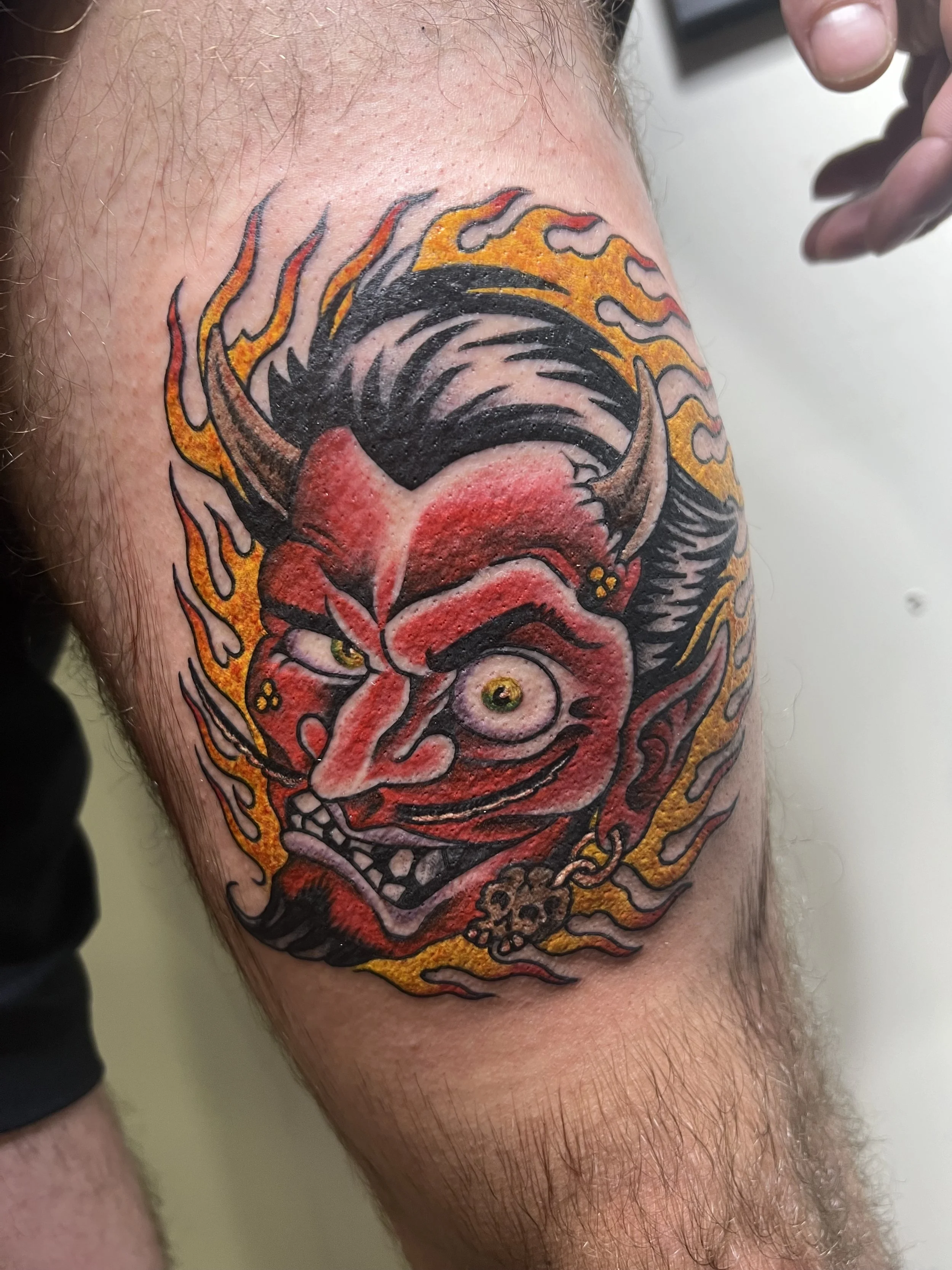 Tattoo of a demonic face with horns, sharp teeth, and intense eyes, surrounded by fiery flames with black, red, yellow, and orange colors, made at Thanks Tattoo