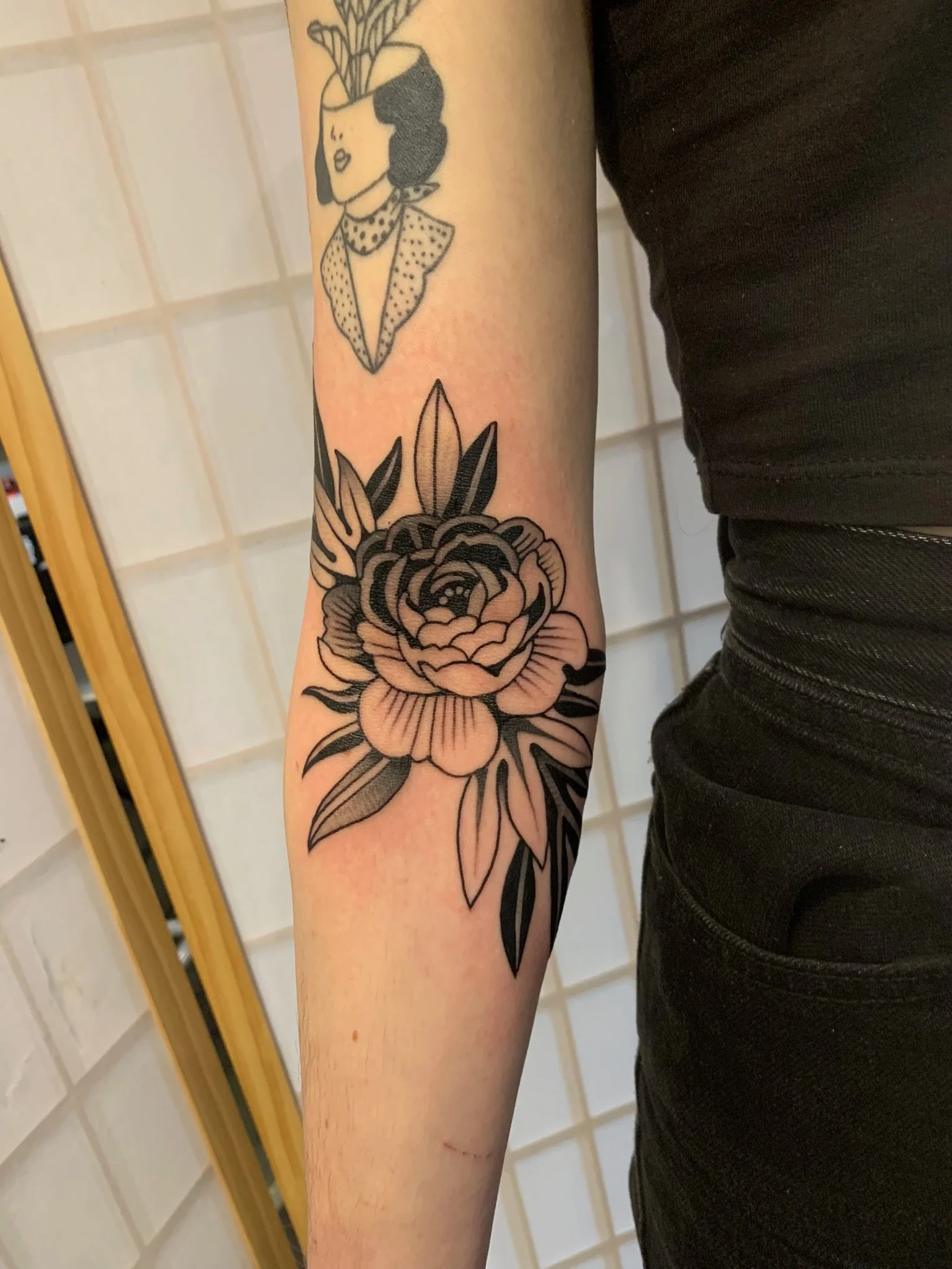 Tattoo of a woman with black hair, wearing a patterned scarf, above a large black and pink peony flower with leaves, on a person's forearm, made at Thanks Tattoo