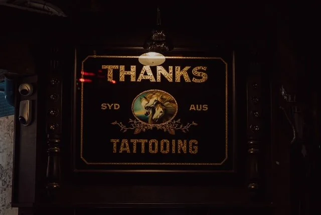 A sign on a dark wall with the words 'Thanks Tattooing' and 'SYD AUS'. The sign features an illustration of a horse and decorative elements, illuminated by a hanging light.