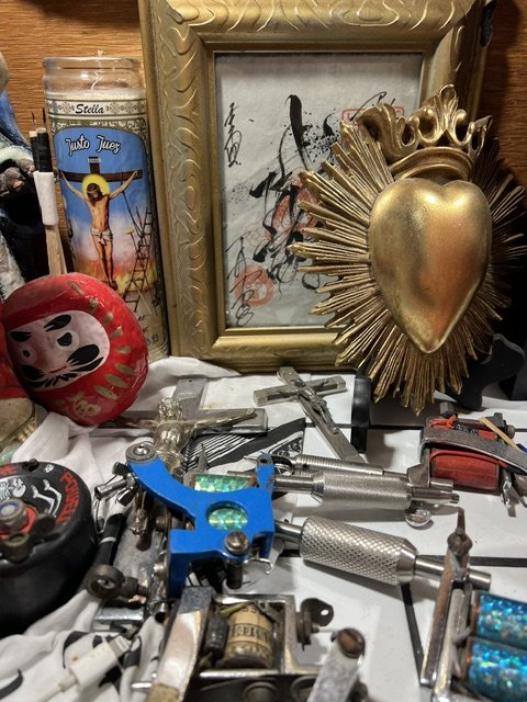 A cluttered surface with religious and decorative items, including a framed picture with graffiti, a golden heart with sun rays, crosses, tools, a Daruma doll, and a candle, in Thanks Tattoo shop