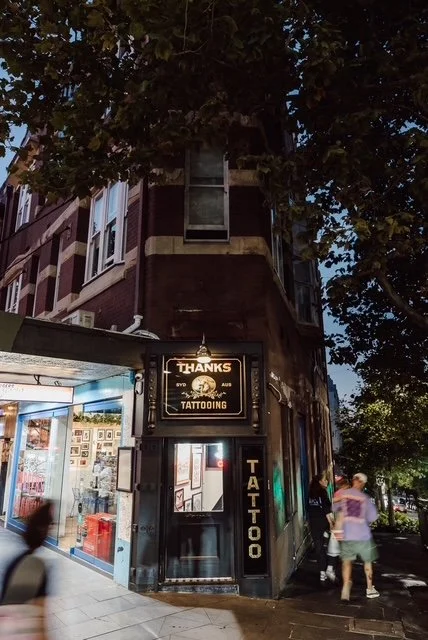 Street corner with tattoo shop sign reading "Thanks Tattooing" and a lit display window, along with pedestrians walking nearby