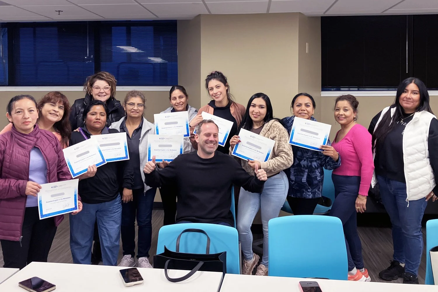 male teacher with thumbs up smiles with his smiling adult students behind him holding certificates of completion, standing within a conference room