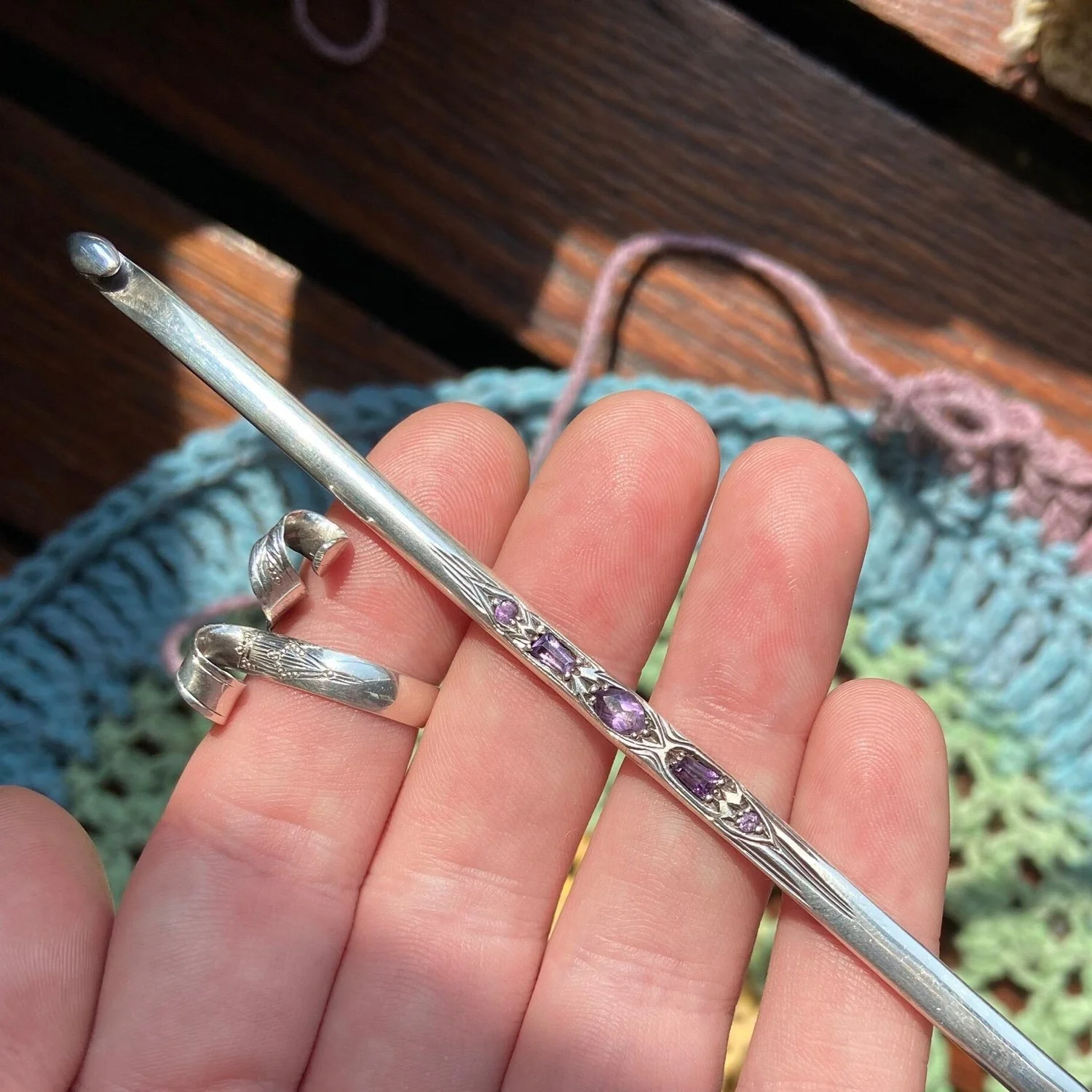 Close-up of a hand holding a silver hairpin with purple gemstones, with rings on fingers and a crocheted cloth in the background.