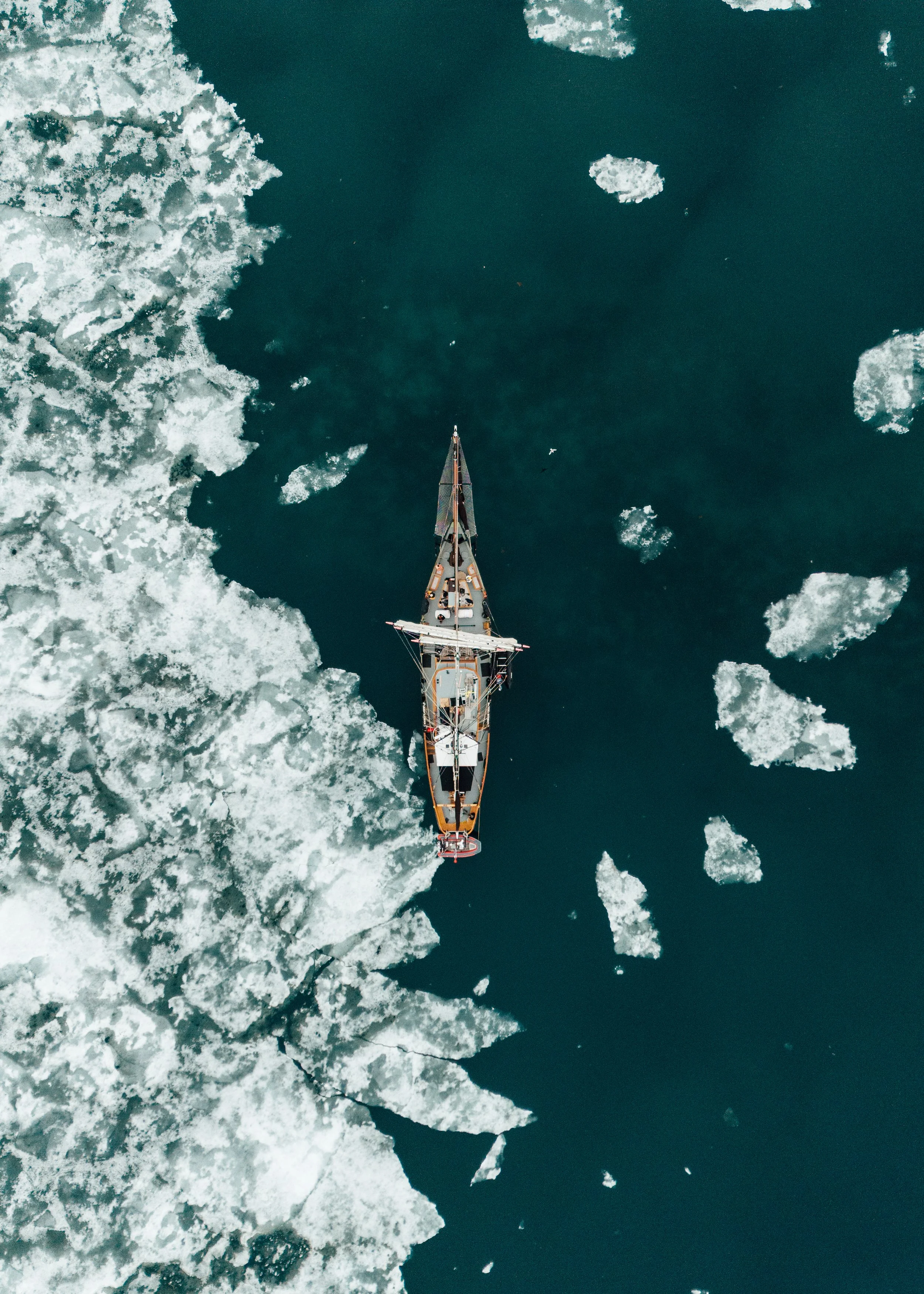 An aerial view of a sailboat navigating through icy waters with ice chunks surrounding it.