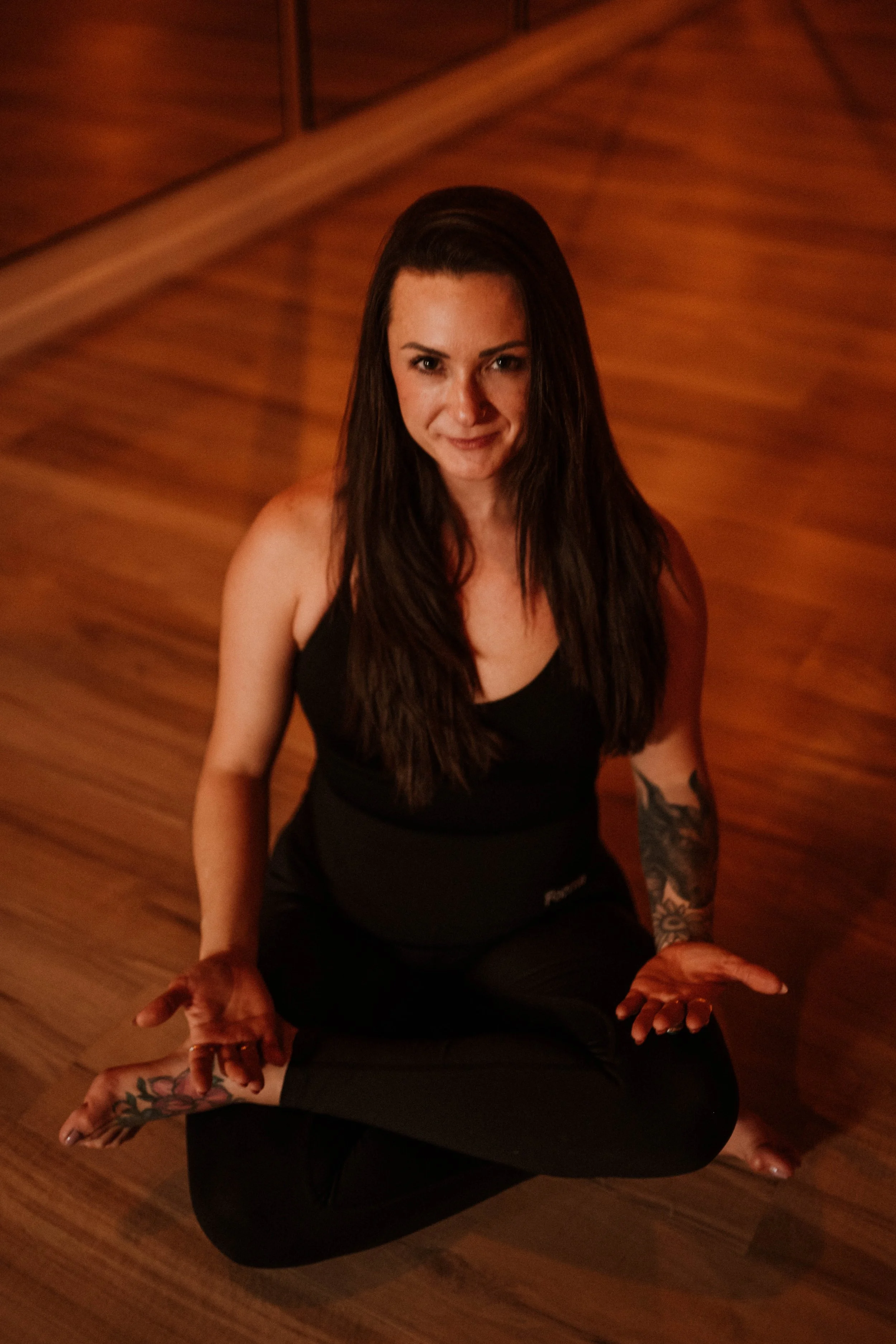 A woman with long dark hair practicing yoga on a wooden floor, sitting cross-legged with hands resting on her knees, wearing a black tank top and black leggings, smiling at the camera.