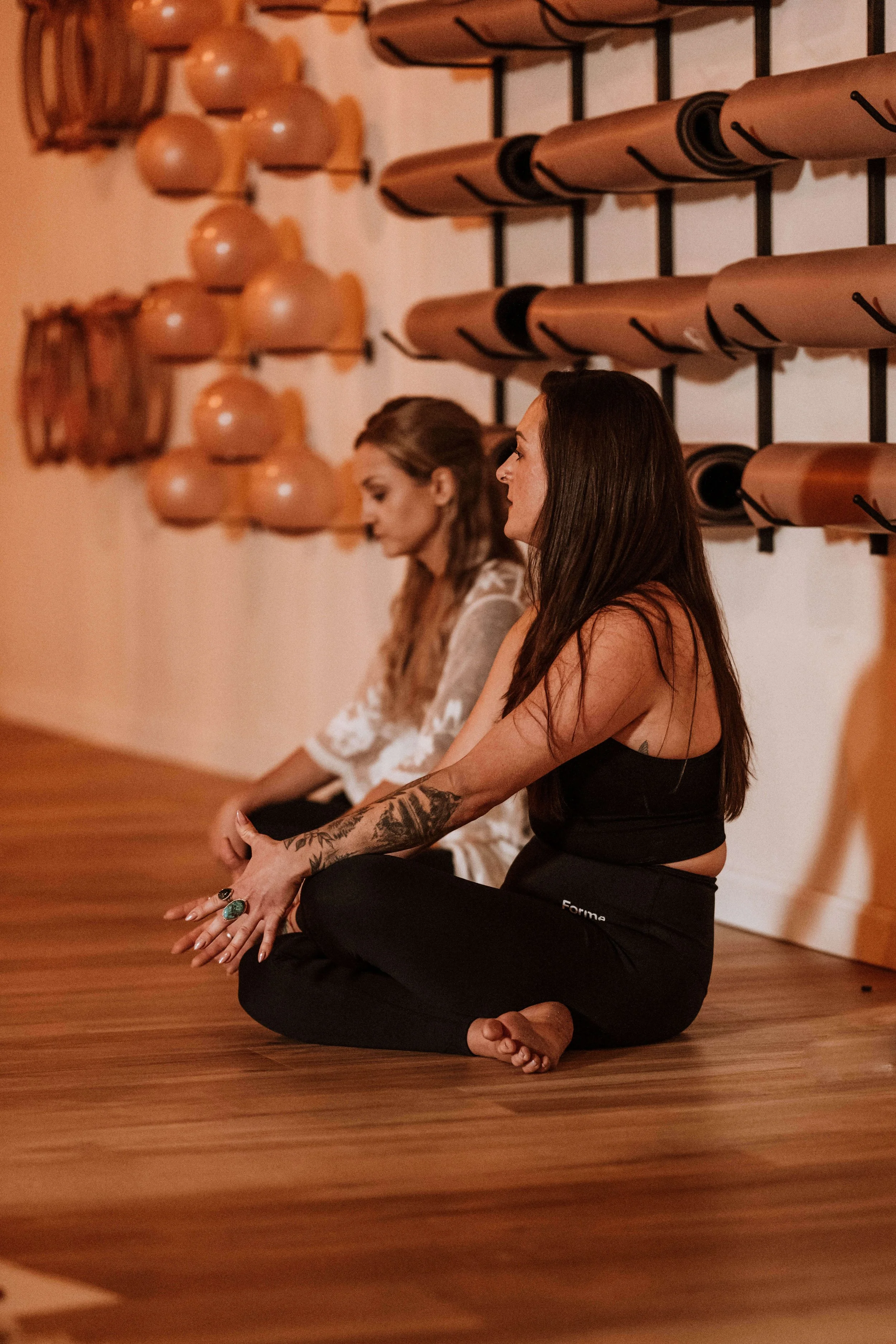 Two women sitting cross-legged on the wooden floor, practicing meditation in a yoga studio with wall-mounted yoga mats and exercise balls.