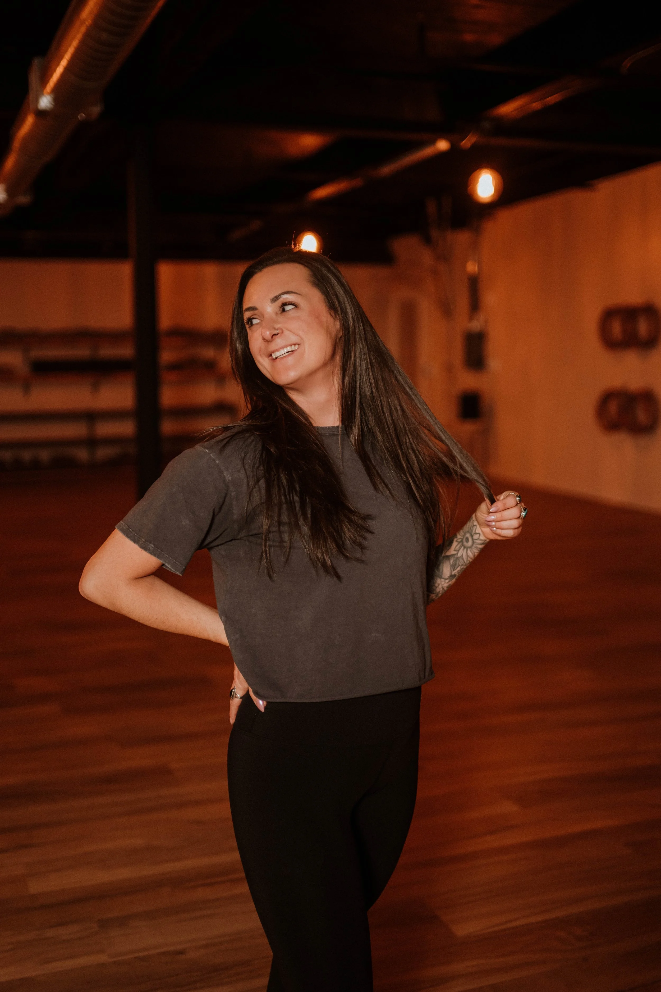 A woman with long dark hair, wearing a gray t-shirt and black pants, standing in a dimly lit dance studio with wooden floors and warm lighting.