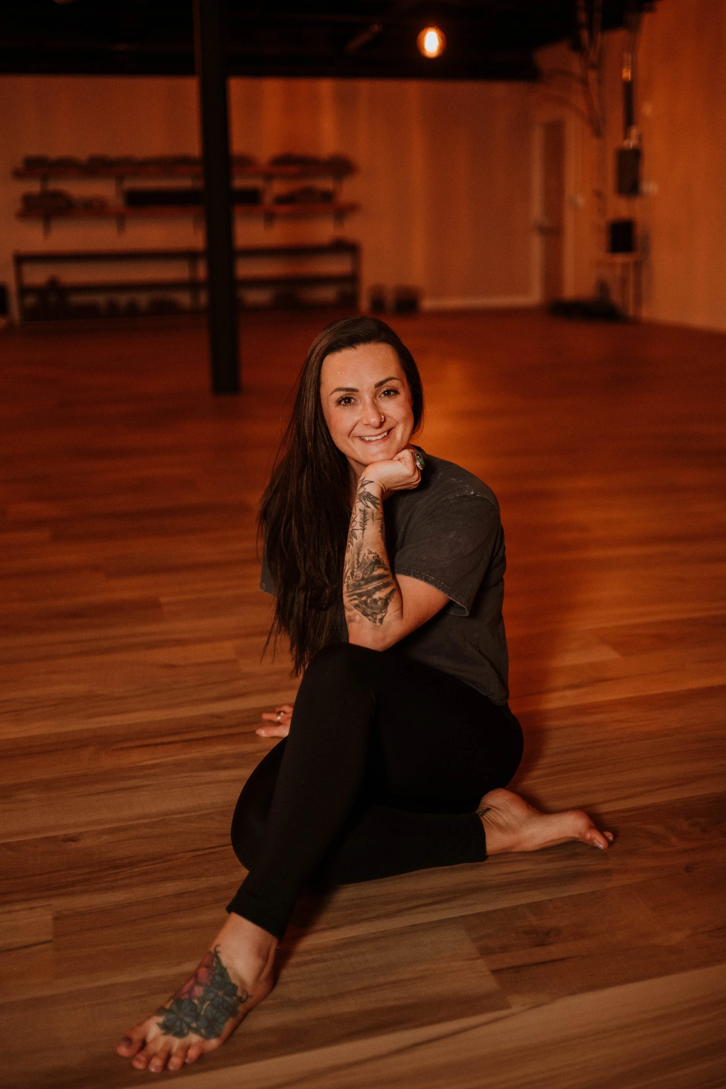 A woman with long dark hair and tattoos sitting on wooden floor in a dimly lit room, smiling at the camera.