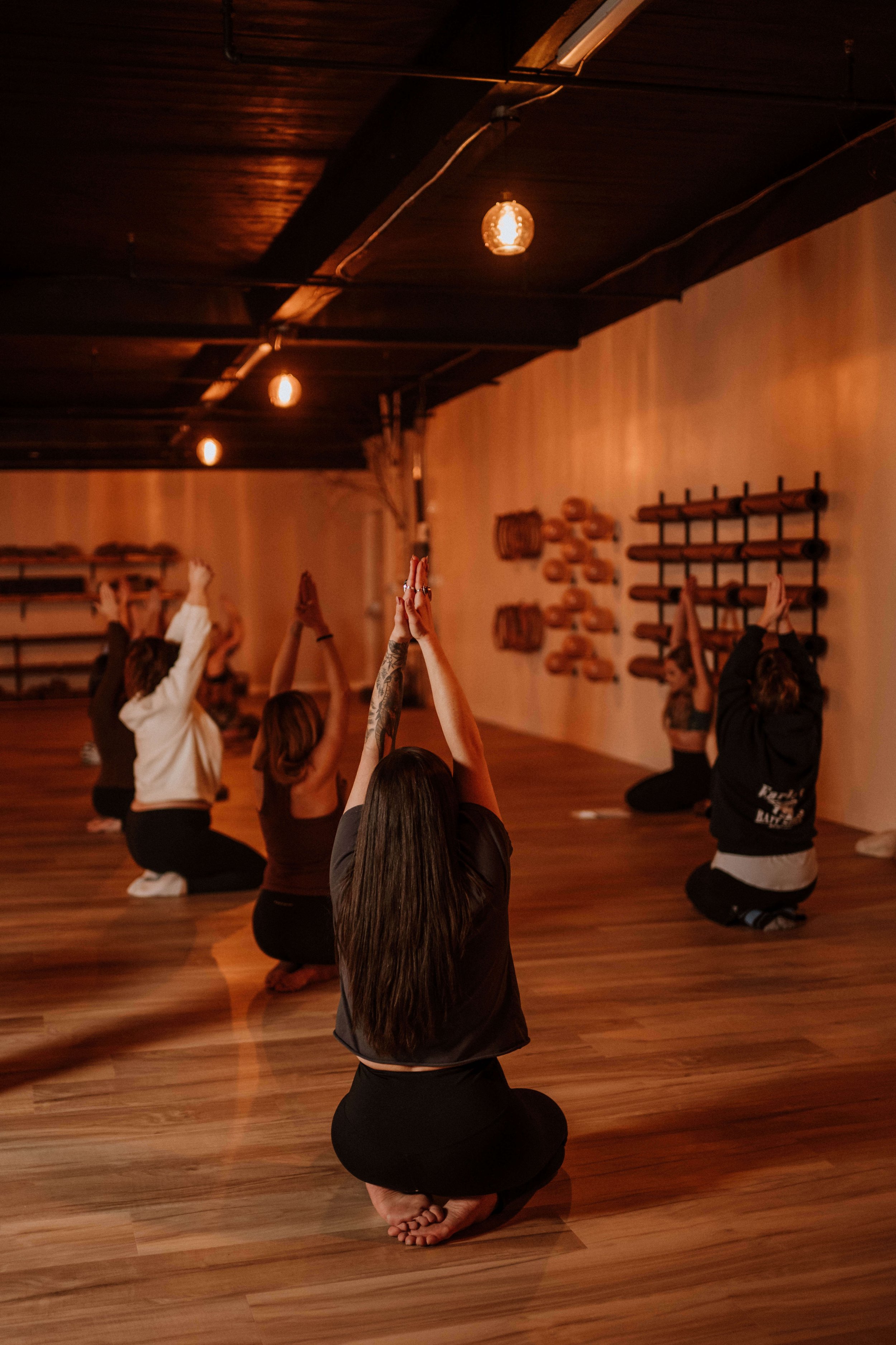 A group of people practicing yoga in a dimly lit studio with wooden flooring and wall decor.