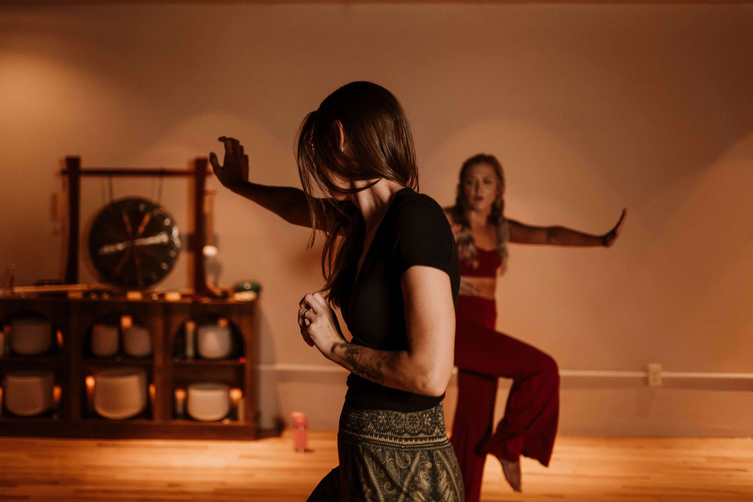 Two women practicing yoga in a dimly lit room, with one in the foreground looking down and the other in the background standing on one leg with arms extended.