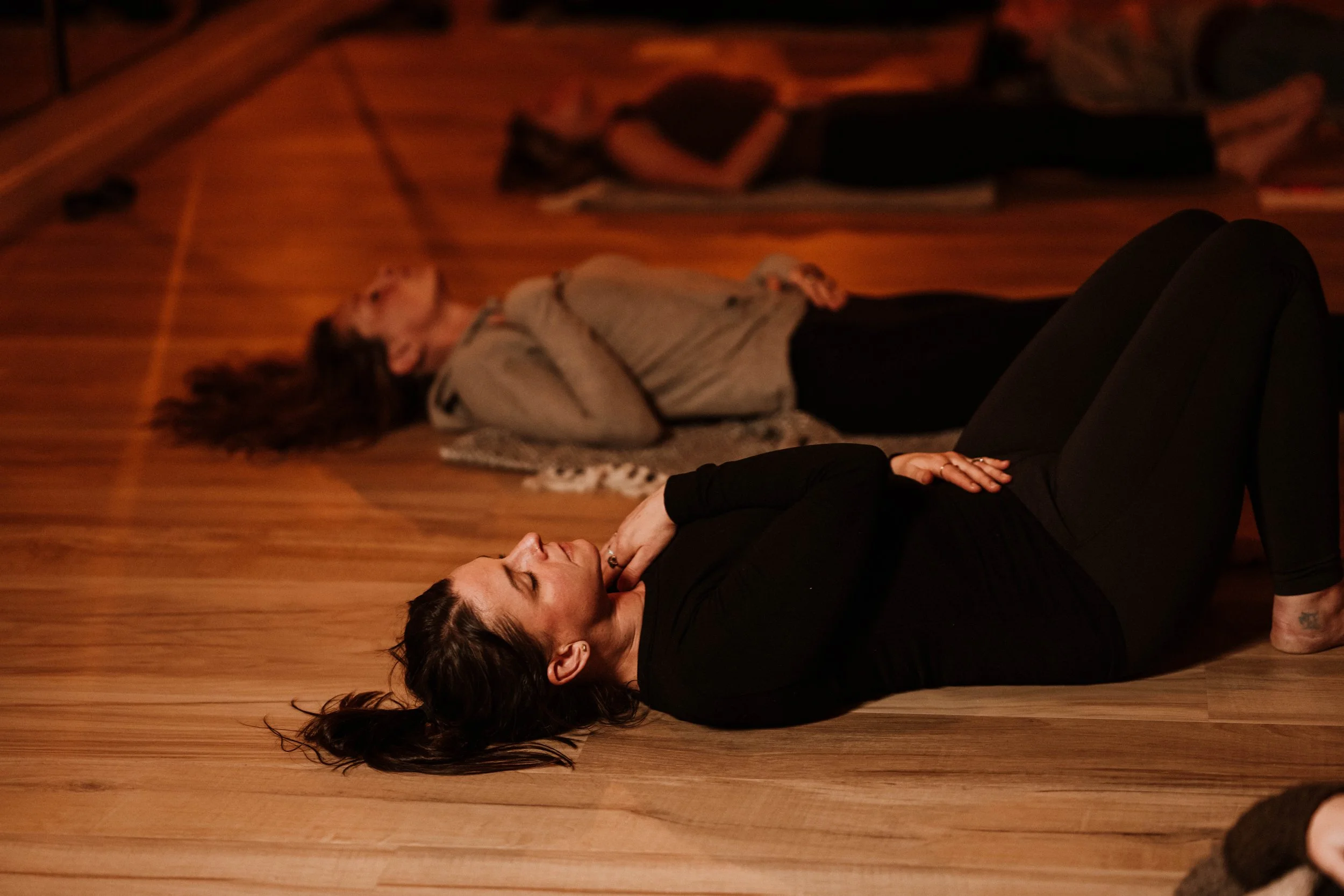 Two women lying on the wooden floor with eyes closed, appearing to meditate or relax in a peaceful setting.