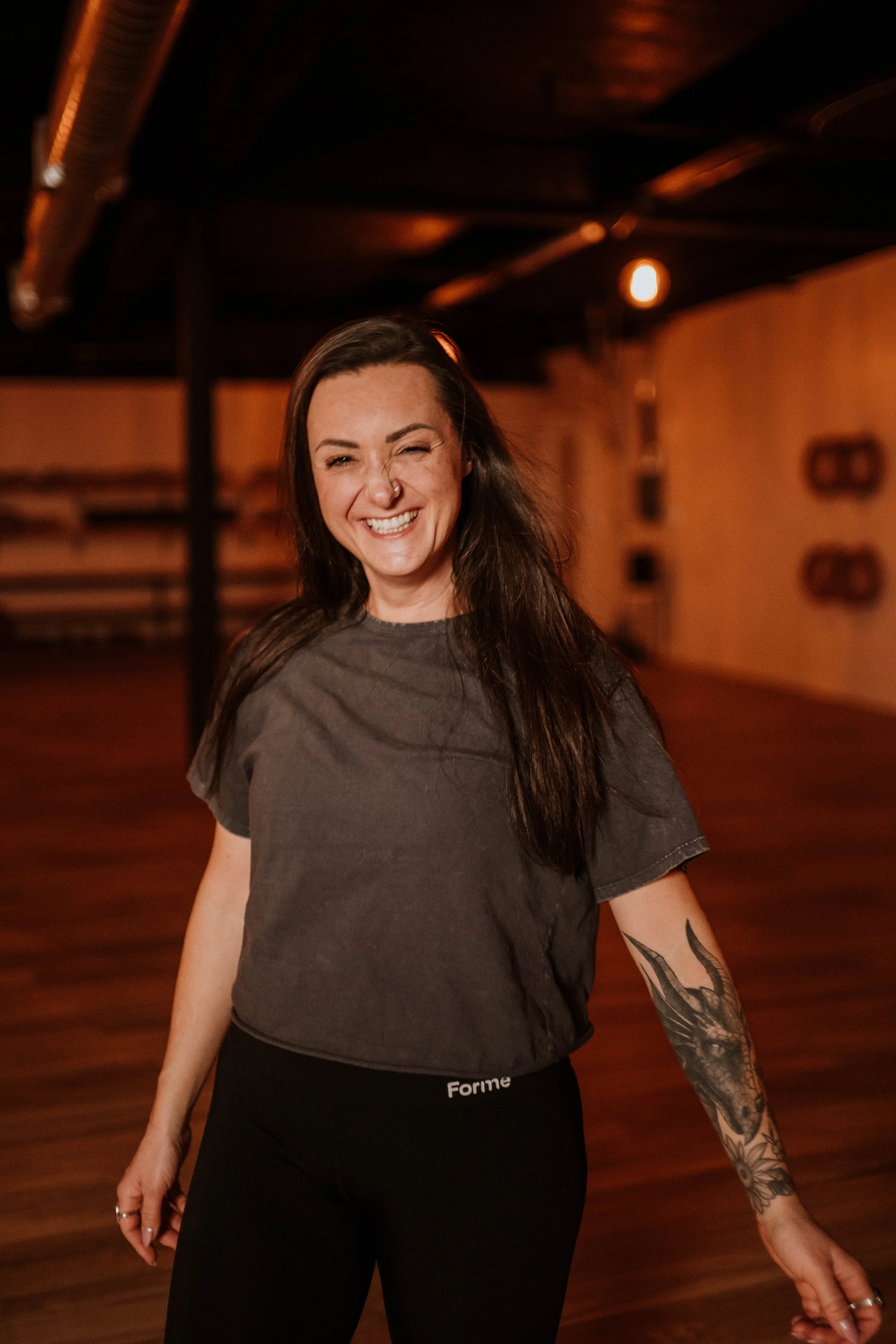 A woman with long dark hair, a nose piercing, and a tattoo on her left forearm, smiling and standing in a dimly lit indoor setting.