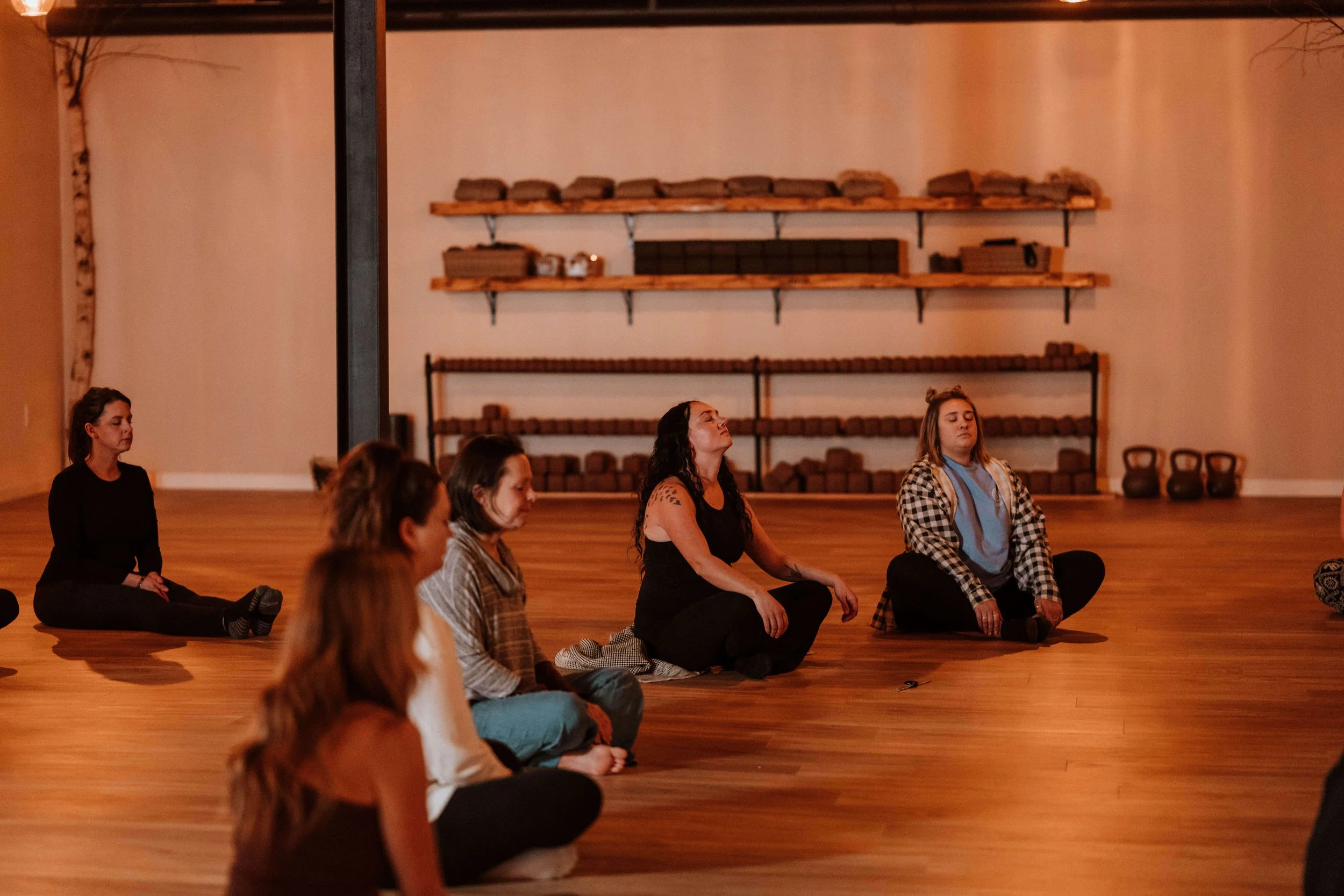 Group of people sitting cross-legged on a wooden floor in a spacious room, practicing meditation or yoga. The room has shelves with weights and kettlebells, indicating it may be a fitness or wellness studio.