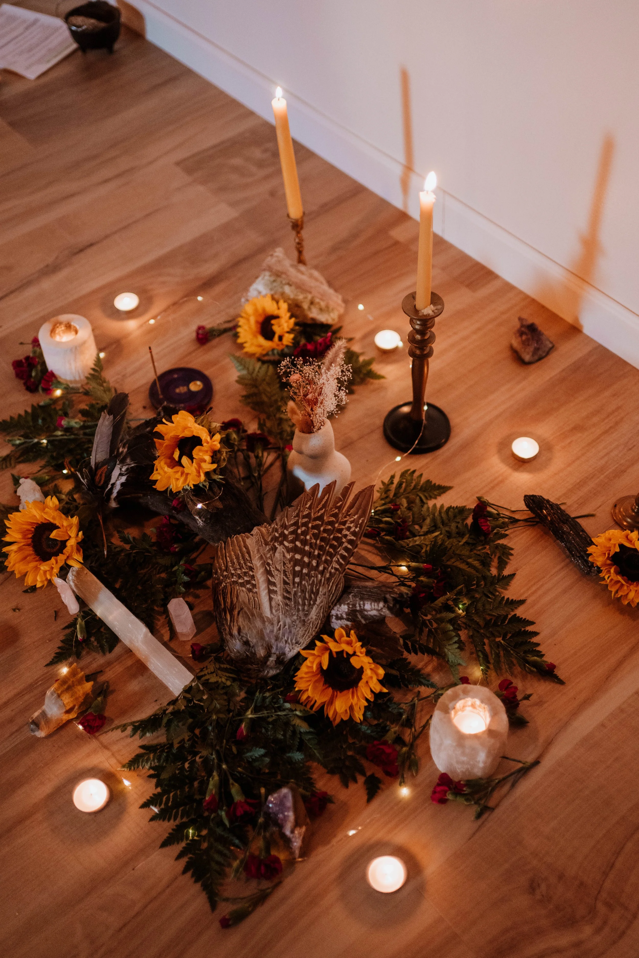 A woodland-themed altar with sunflowers, feathers, candles, crystals, and natural decorations arranged on a wooden floor.