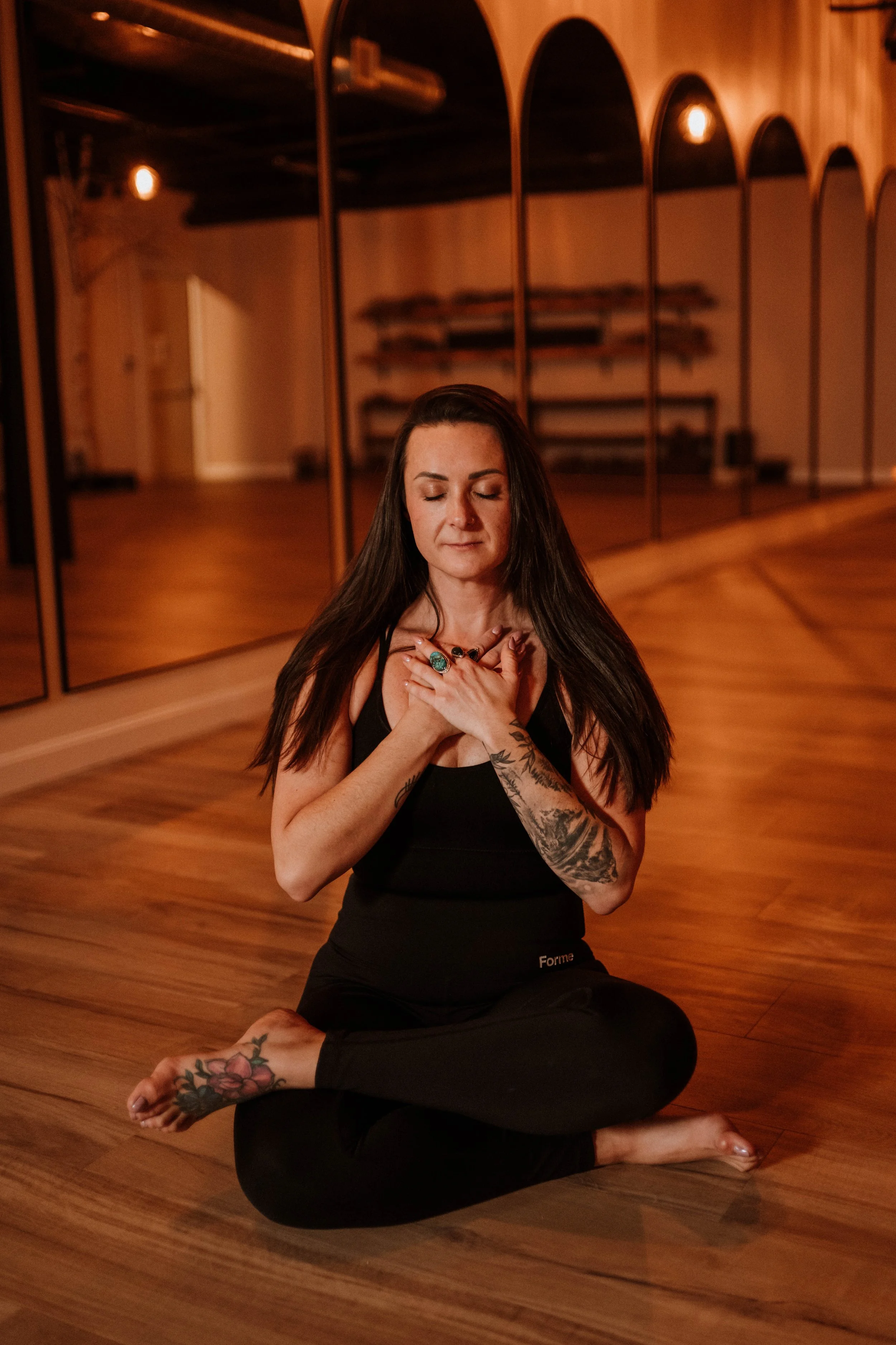 A woman practicing yoga in a dance studio, sitting cross-legged on the wooden floor with her eyes closed, hands over her chest, wearing black athletic wear, and tattoos on her arms and feet.