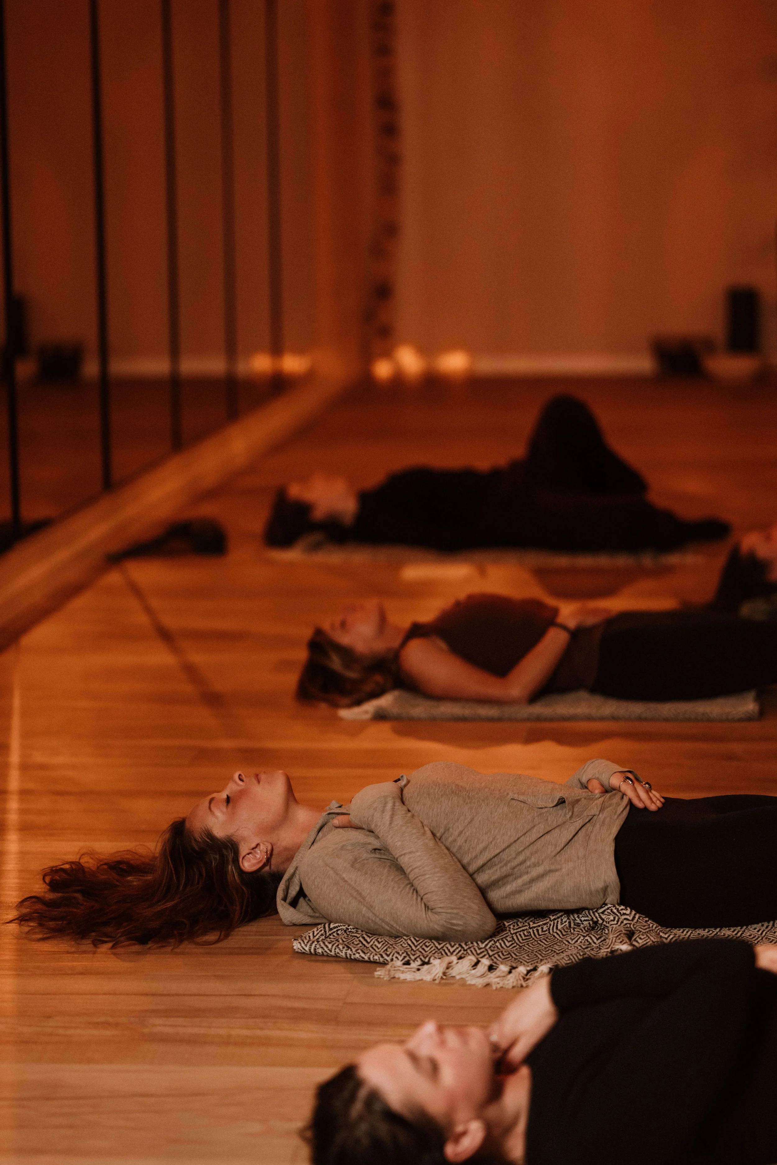 Women practicing yoga or meditation on mats in a dimly lit studio with a wooden floor.