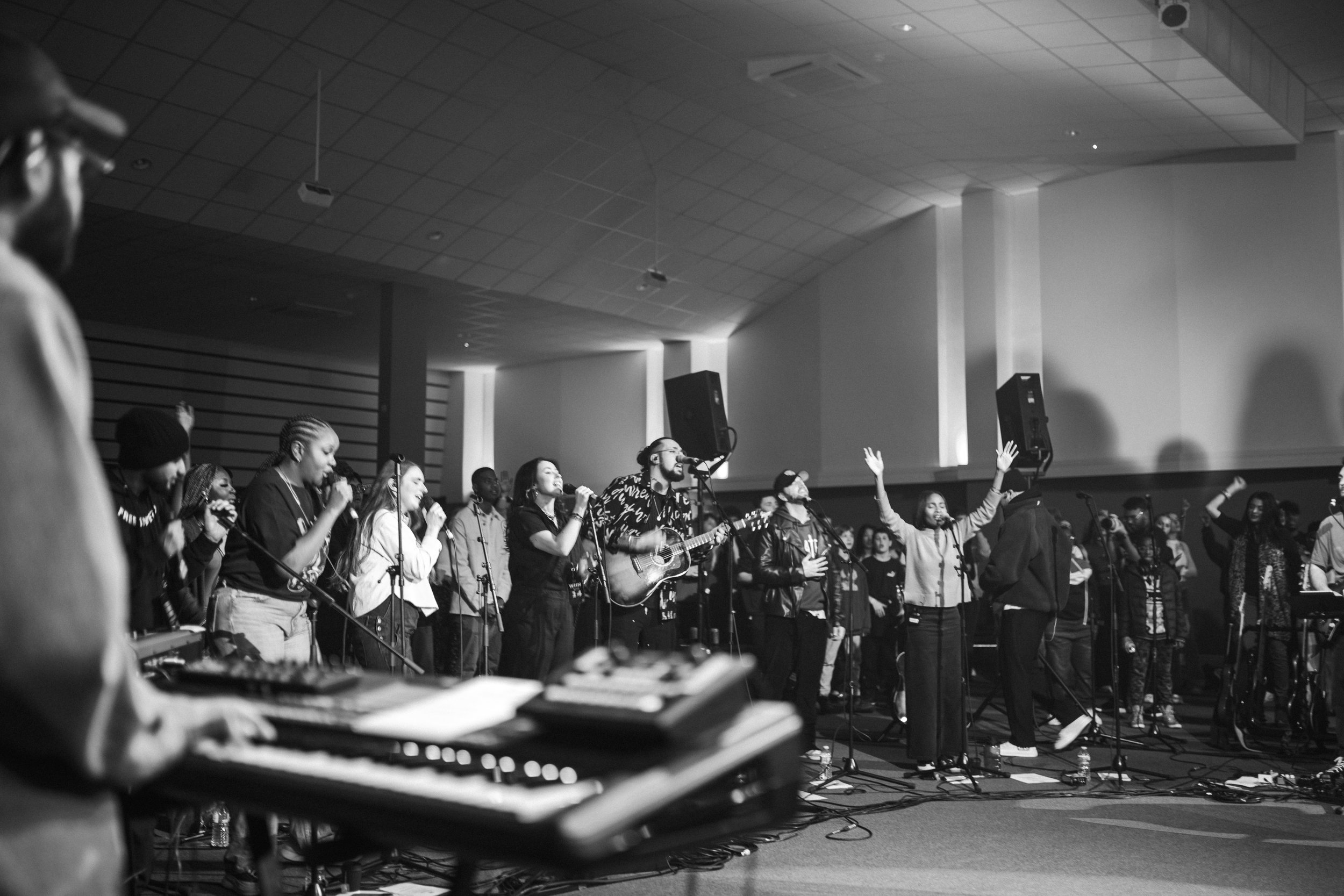A group of musicians and singers performing on stage in a black and white image. Some are playing instruments like a guitar and keyboard, while others sing into microphones. The audience is visible in the background, with people raising their hands.
