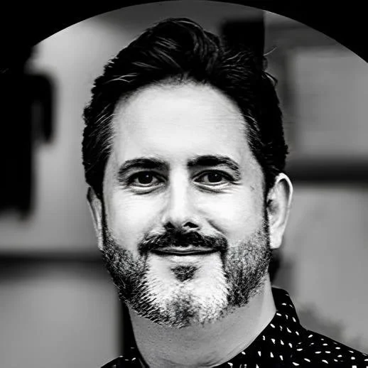 Black and white portrait of a man with dark hair, a beard, and mustache, wearing a dark, patterned shirt, smiling slightly, in an indoor setting.