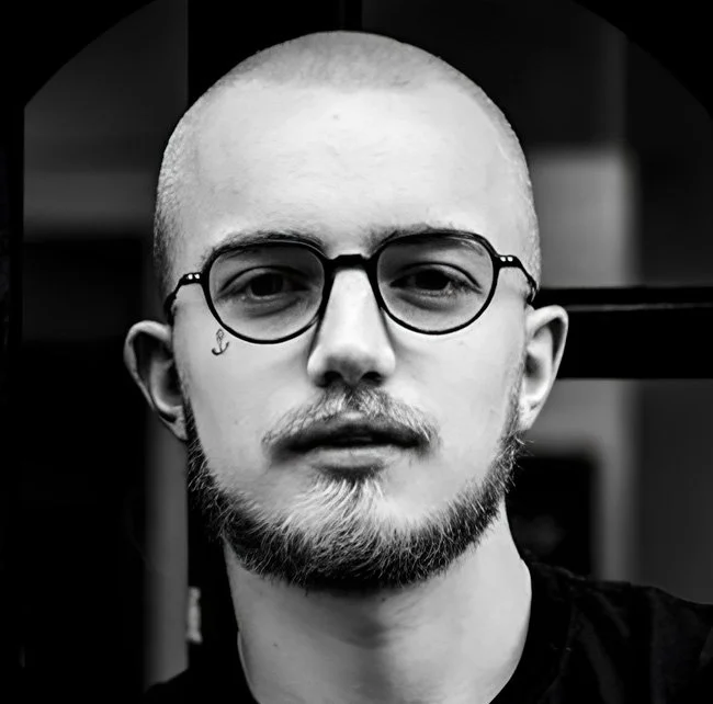 A young man with a shaved head, glasses, and a beard, looking directly at the camera with a neutral expression, in a monochrome setting.