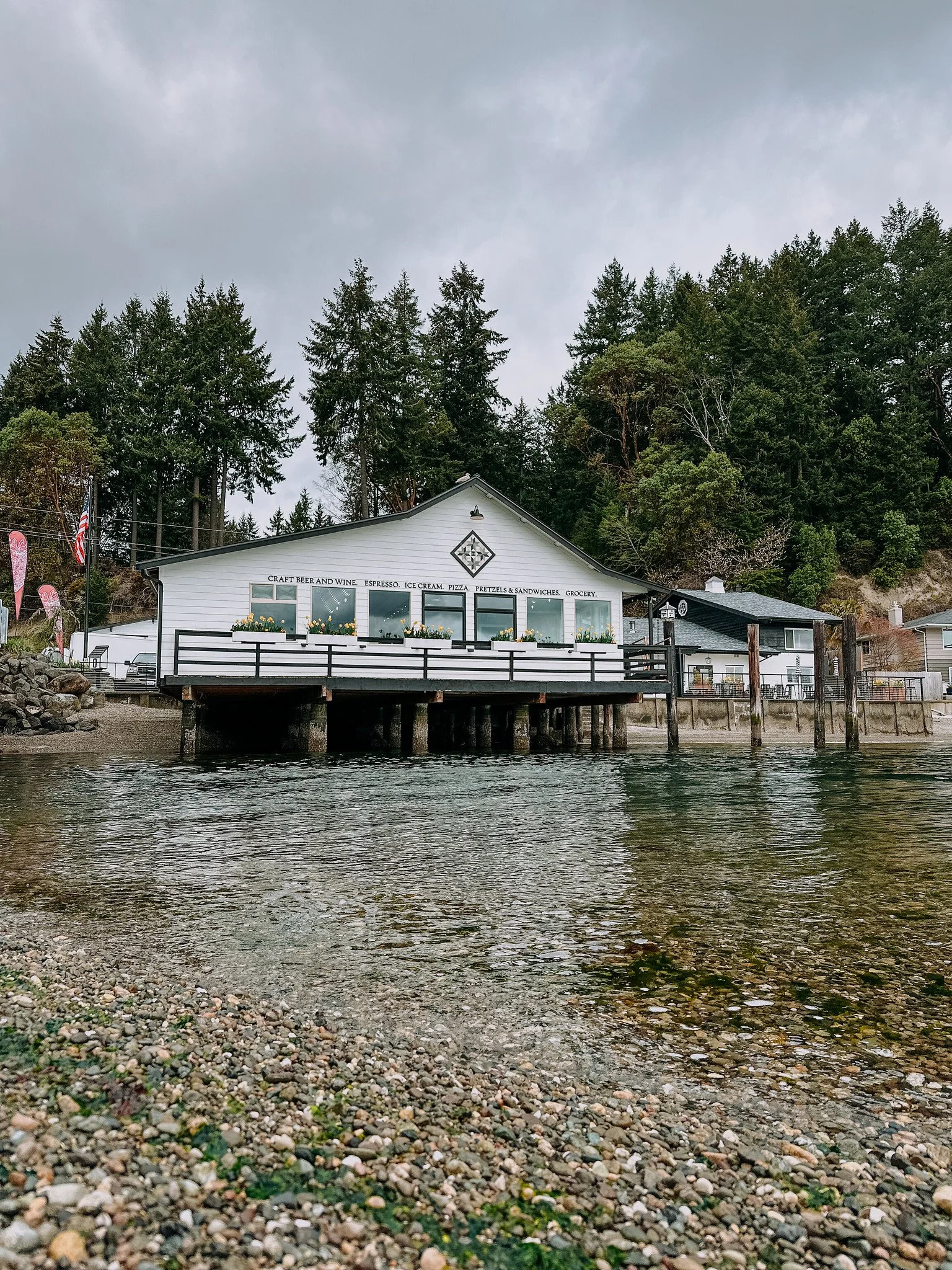 Olalla Bay Market and Landing with outdoor seating, signage listing craft beer, wine, espresso, ice cream, pizza, pretzels, sandwiches, and groceries, surrounded by trees under a cloudy sky.