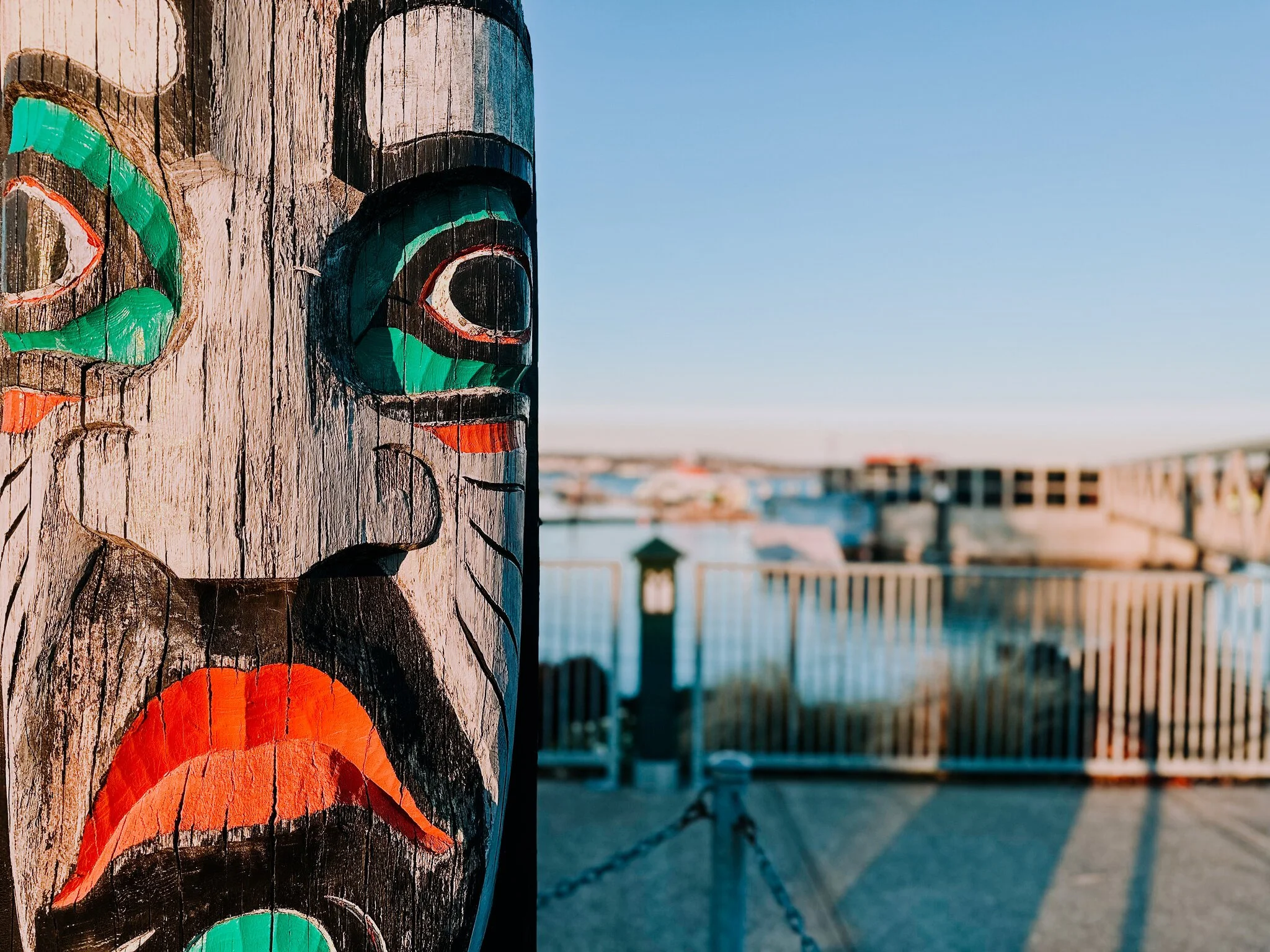 Close-up of a carved wooden totem pole with colorful paint, featuring a face with prominent eyes, nose, and mouth, set against a blurred outdoor waterfront scene with a bridge and blue sky on the Port orchard waterfront.