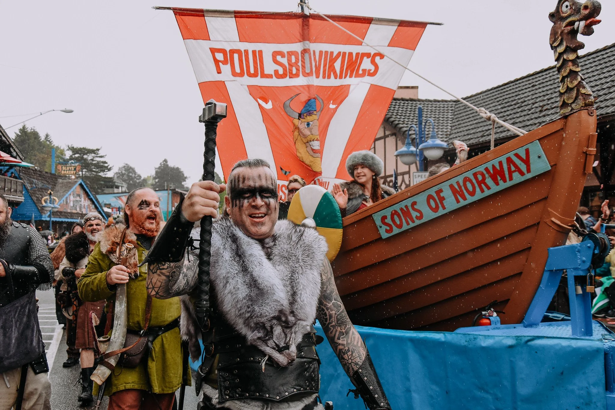 A group of people dressed in Norse and Viking costumes participating in a parade or festival, with a float decorated as a Viking ship labeled "Sons of Norway" and a banner reading "Poulsbovings" in the background. The main person in the foreground is smiling and holding a staff.