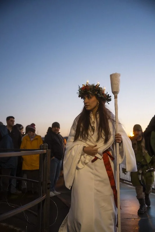 Young woman dressed in ceremonial white robes with a red sash, wearing a flower crown, holding a torch, participating in Julefest in Poulsbo at sunset with onlookers in the background.