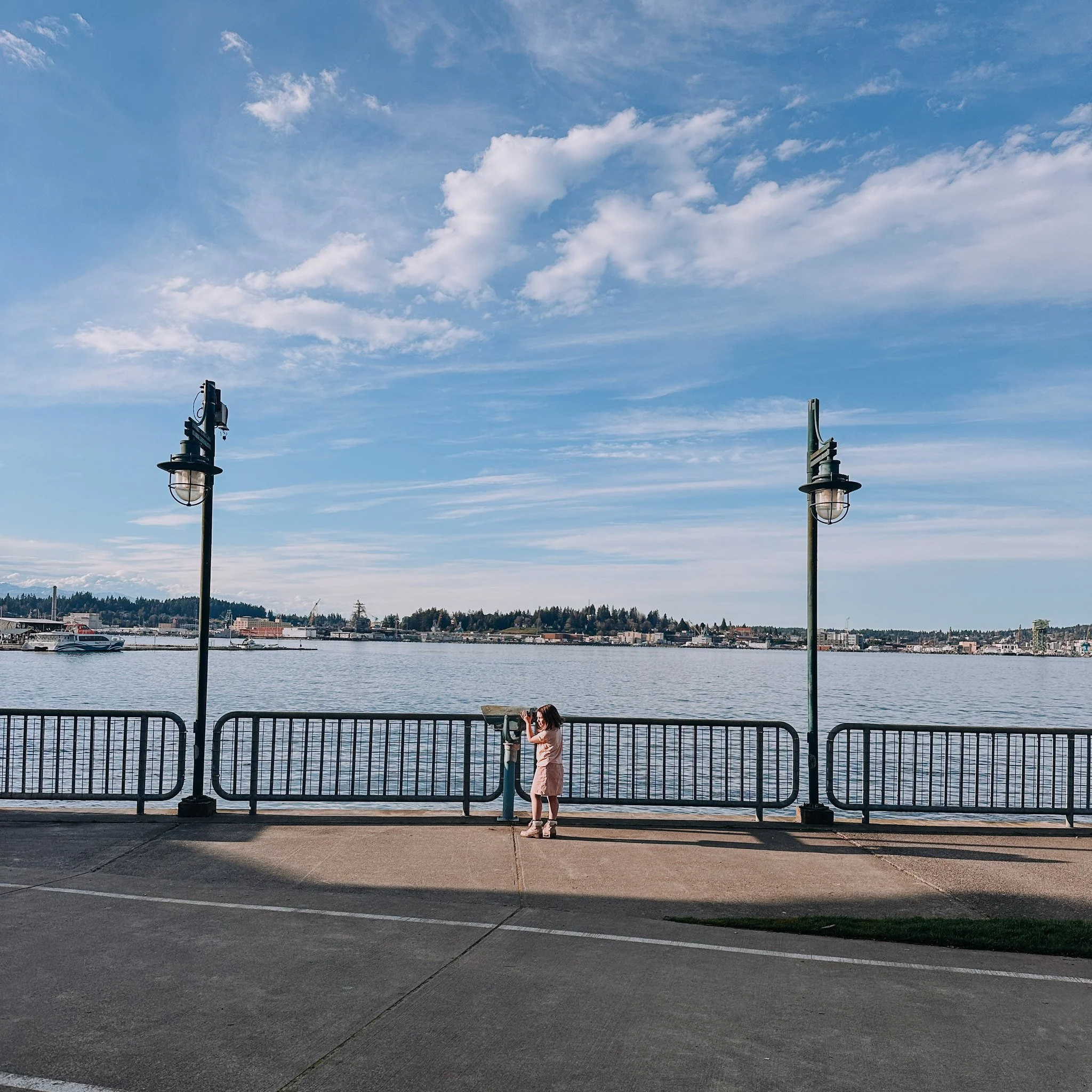 A child standing on a sidewalk next to a railing by the water, looking through a telescope mounted on the railing, with a view of a lake, trees, and buildings in the background under a partly cloudy sky on the Port Orchard waterfront. 