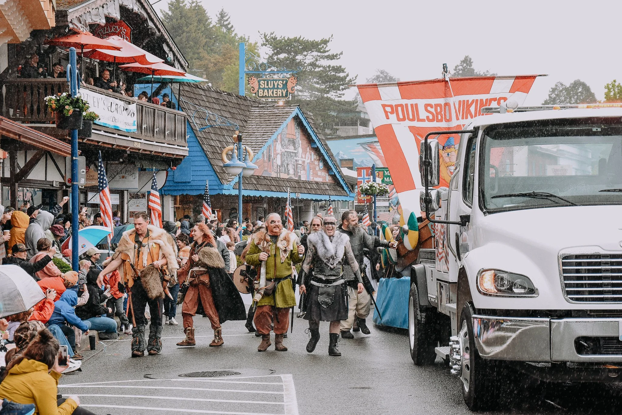 People dressed in Viking costumes parading on a street during the Vikingfest festival in Poulsbo with onlookers sitting and standing, some taking photos, under umbrellas in rainy weather.