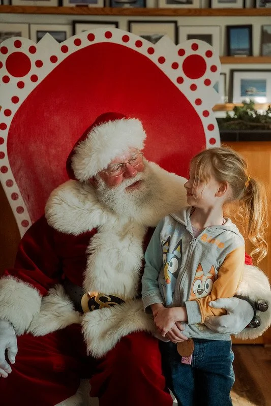 A young girl smiling and looking at Santa Claus, who is sitting on a red and white throne, wearing a traditional red suit with white fur trim, in a festive setting.