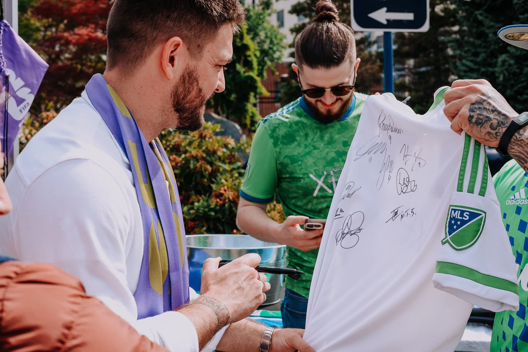 Two men are signing and holding soccer jerseys outdoors, with trees and a street sign in the background. One man is wearing a white shirt with a purple and yellow scarf, and the other is in a bright green shirt with sunglasses.