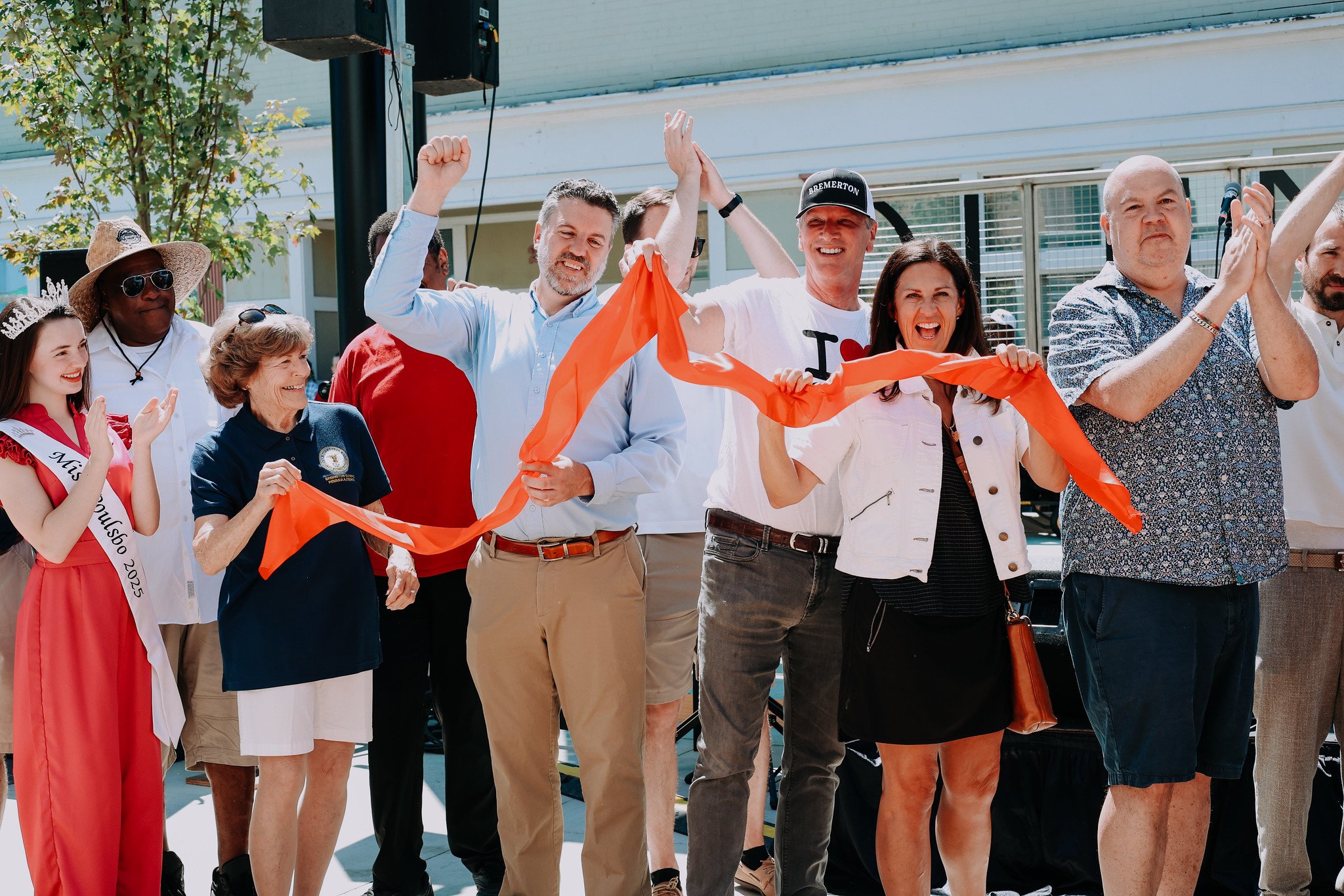 Group of people celebrating a ribbon-cutting ceremony for Quincy Square outdoors on a sunny day. They are smiling, clapping, and raising their hands in victory. Some are holding a long orange ribbon.