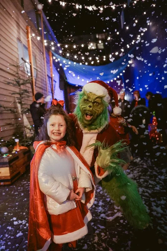 A young girl standing next to a person dressed as the Grinch with green fur, a Santa hat, and a red costume, at Holiday on the Bay Christmas festival in Port Orchard with snow and holiday decorations.