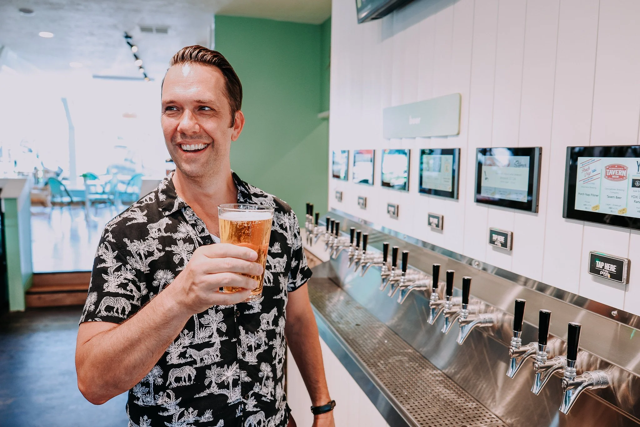 A man with dark hair in a patterned short sleeve shirt holding a glass of beer at a self-serve beer tap station at The Sip in Poulsbo.