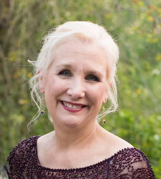 A smiling woman with light blonde hair in an outdoor setting with trees and greenery in the background.
