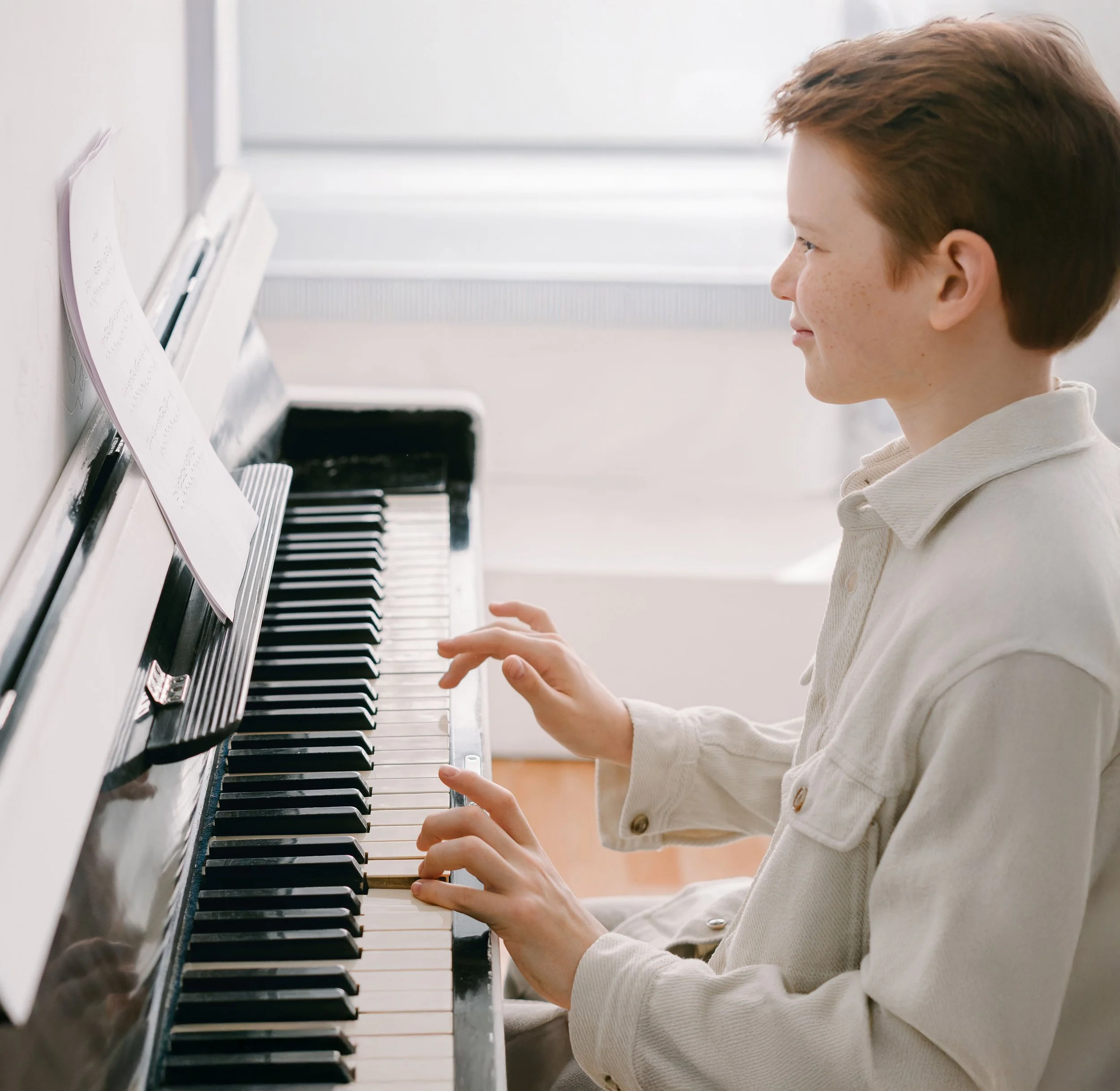 A young person with short brown hair playing a white upright piano, wearing a cream-colored jacket, in a well-lit room with a window in the background.