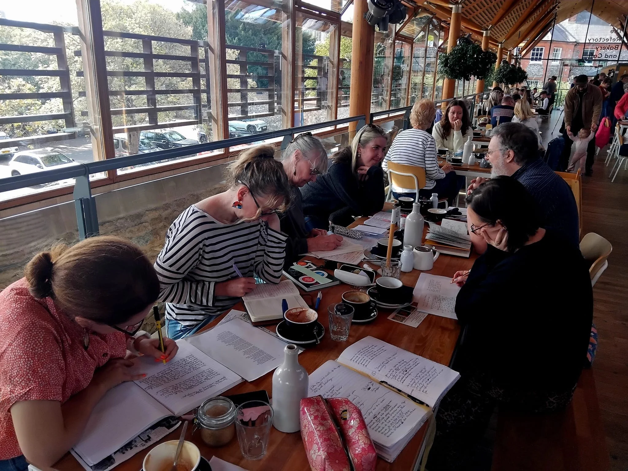 Members of Norwich Writing Teachers group are sat around a rectangular table, heads bowed, writing in notebooks from sheets of prompts placed in front of them.