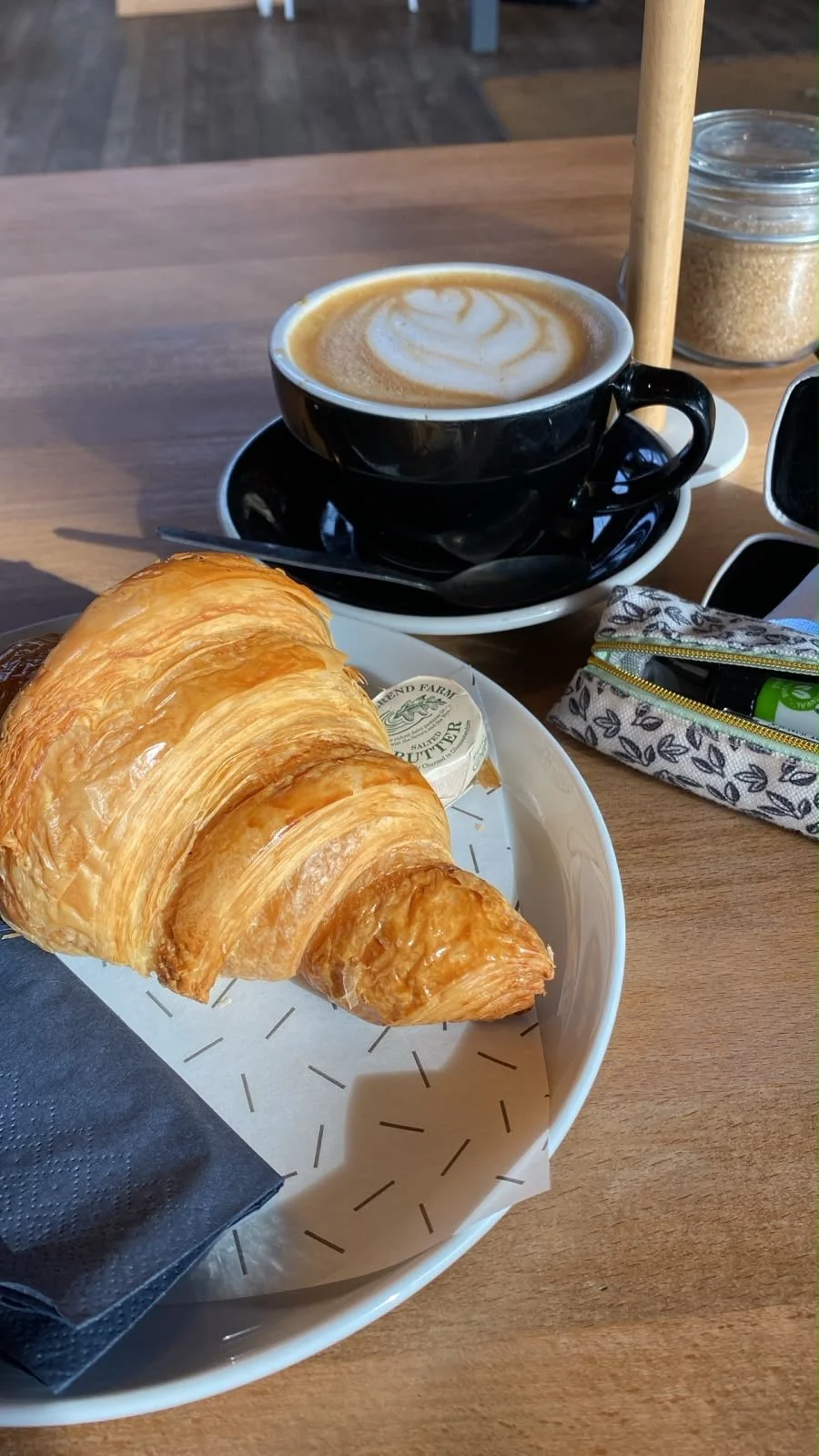 Picture of a latte in a black mug with a heart-motif in the cream, and a croissant on the table.
