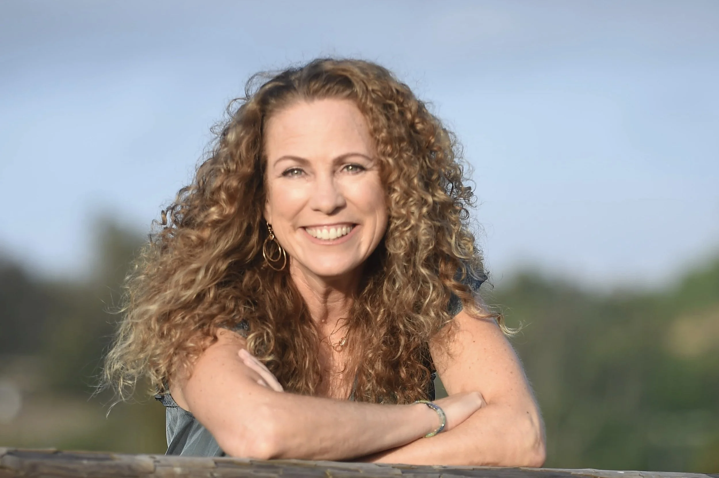 Jeanine Mouchawar, parenting coach for parents of teenagers, smiling and leaning on a wooden fence outdoors, representing calm, emotional support, and parent-child connection.