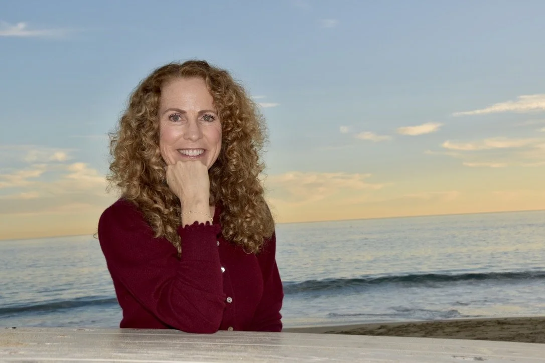 Parenting coach Jeanine Mouchawar sitting at a beach picnic table at sunset, smiling warmly at the camera