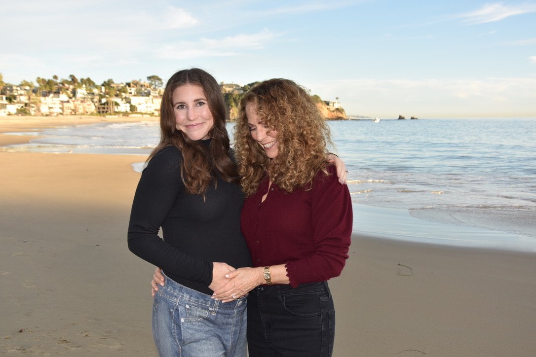 A mother and her adult daughter laughing together on the beach, arms around each other, with the ocean behind them