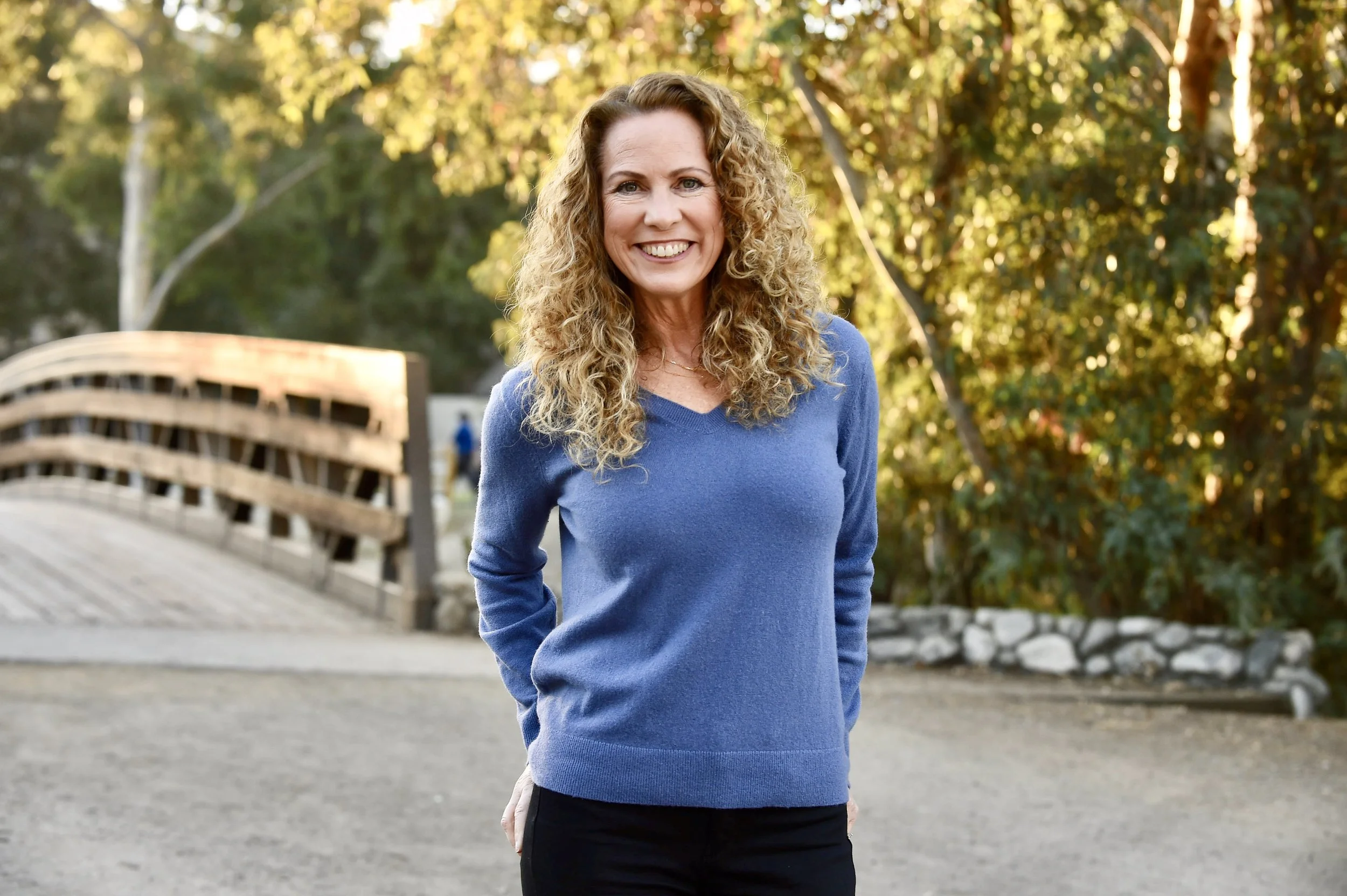 Jeanine Mouchawar, parenting coach for parents of teenagers, smiling in a blue sweater outdoors on a wooden bridge.