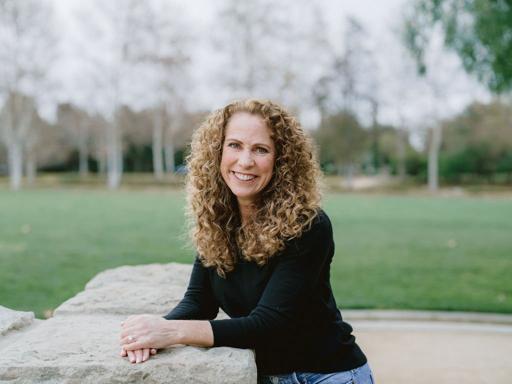 Parenting coach Jeanine Mouchawar leaning on a stone wall outdoors, smiling — representing approachable support for parents of teenagers who feel stuck in repeating arguments and patterns