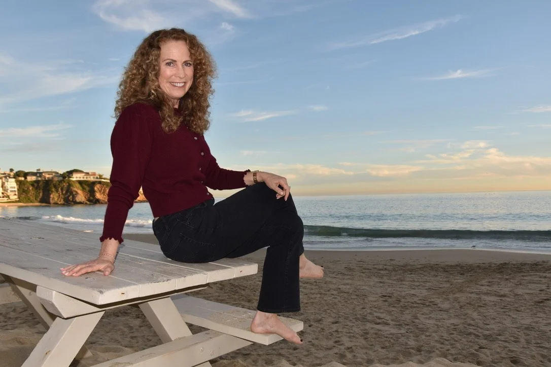 Parenting coach Jeanine Mouchawar sitting at a beach picnic table at sunset, smiling confidently at the camera